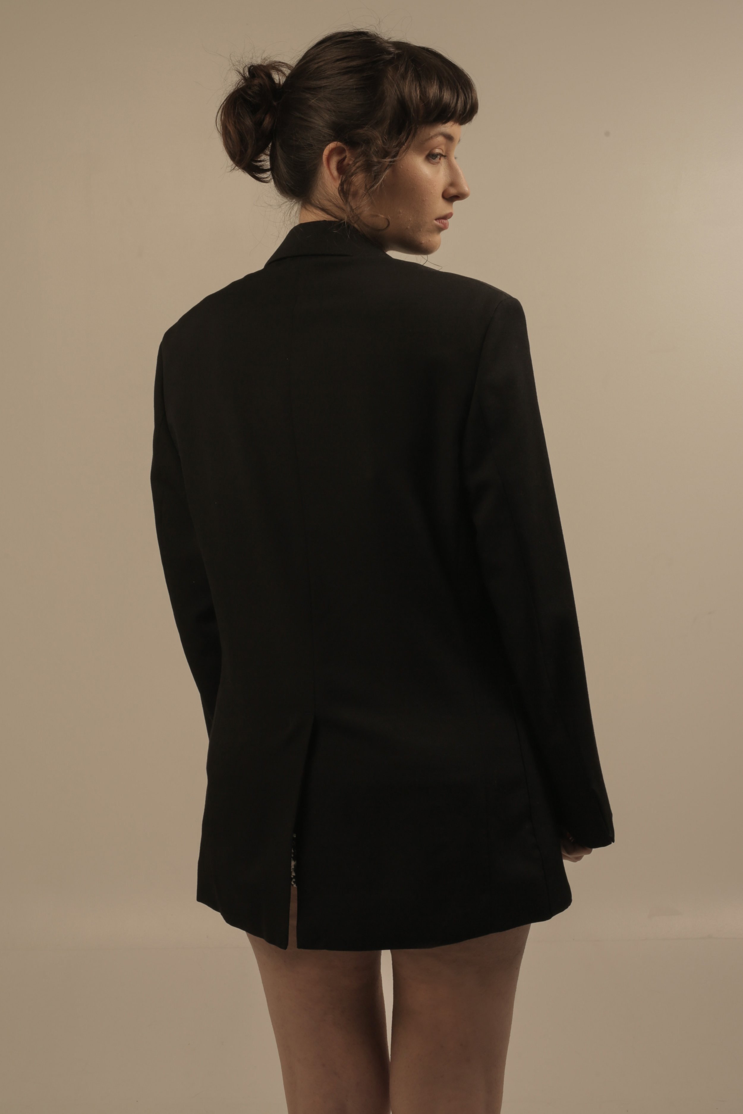 A woman with brown hair in a messy bun, wearing a black blazer, standing against a plain beige wall, looking to the side.