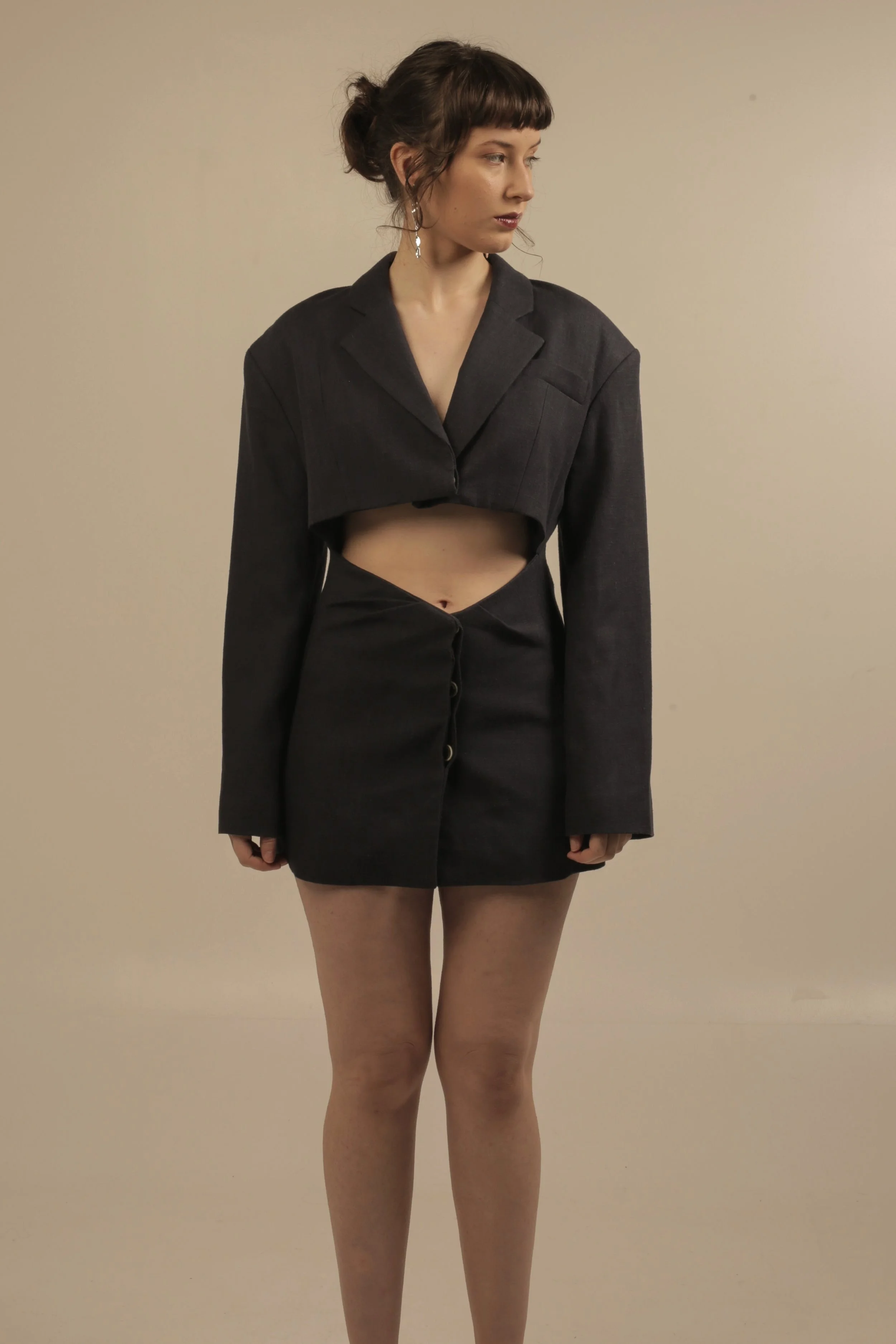 Young woman wearing a cropped dark blazer and high-waisted skirt with buttons, standing against a plain beige wall.