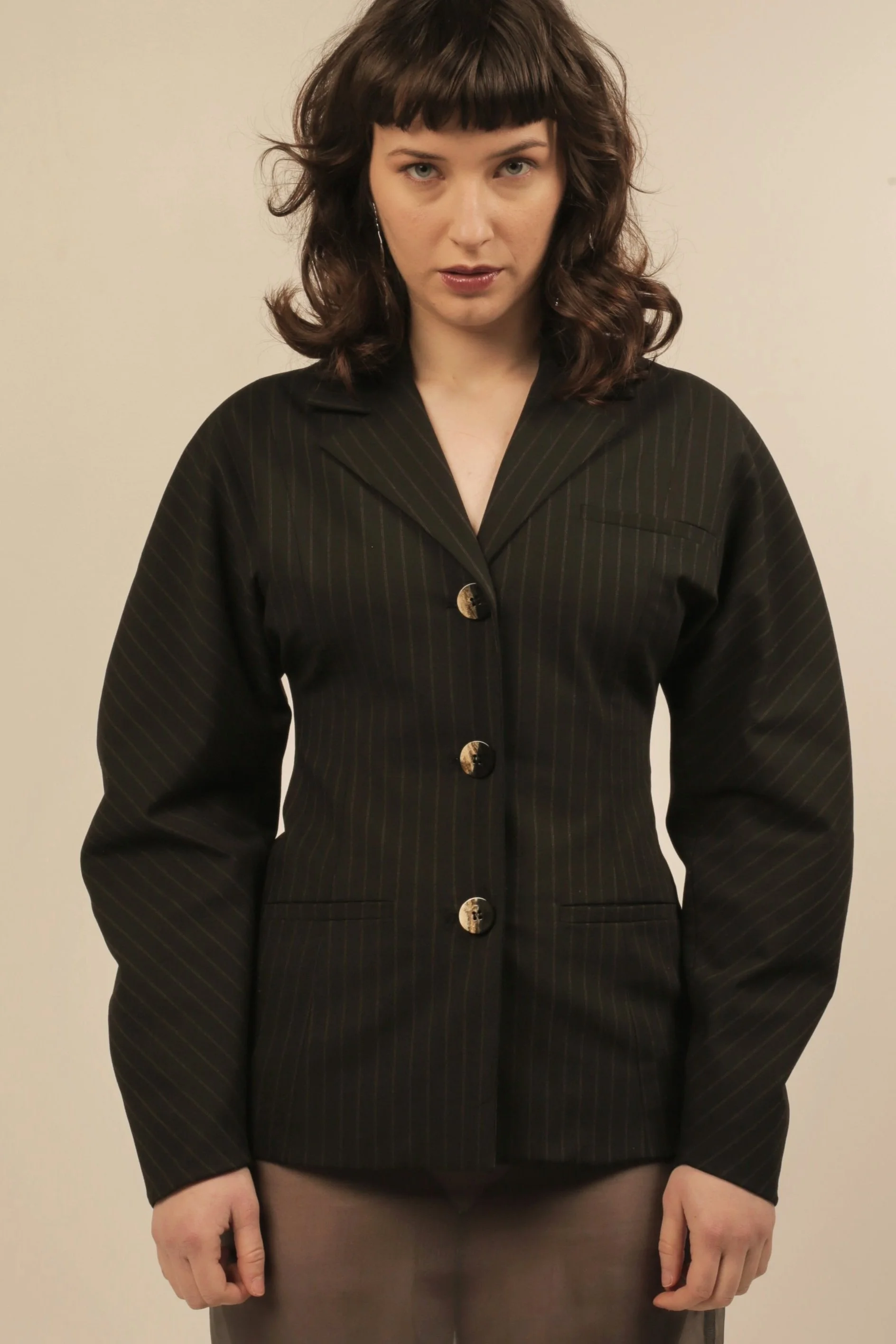 A woman with shoulder-length wavy brown hair, wearing a black pinstripe blazer with gold buttons, standing against a plain beige wall.