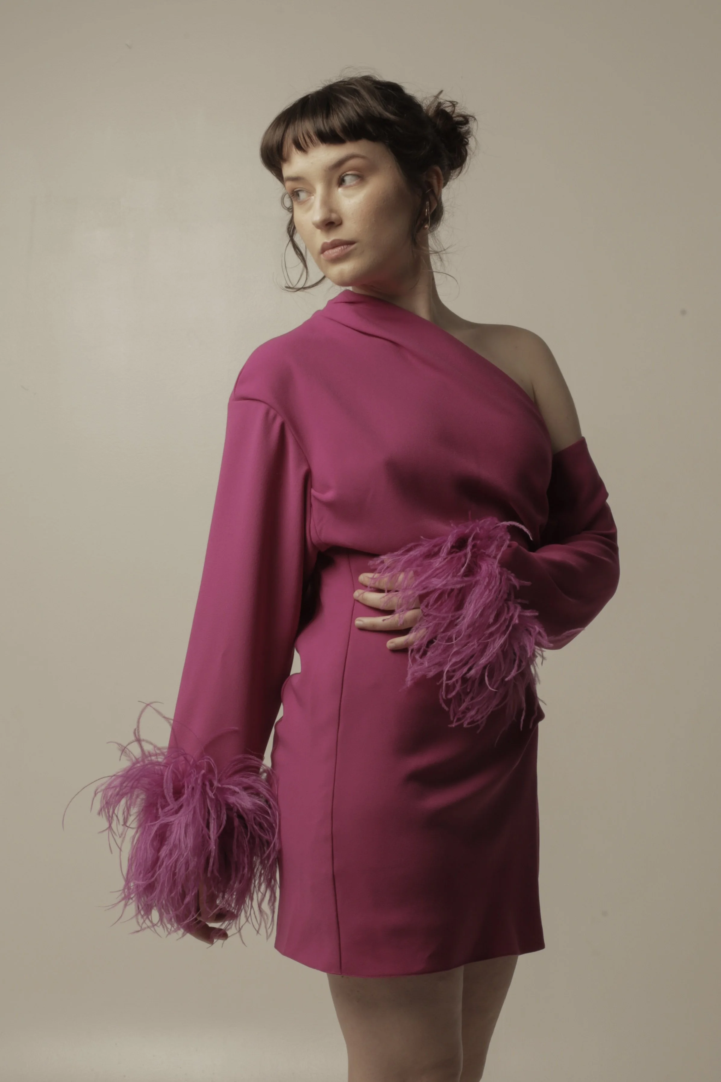 A woman in a pink satin dress with feathered cuffs stands against a plain beige background, looking to her left with her hand on her waist.
