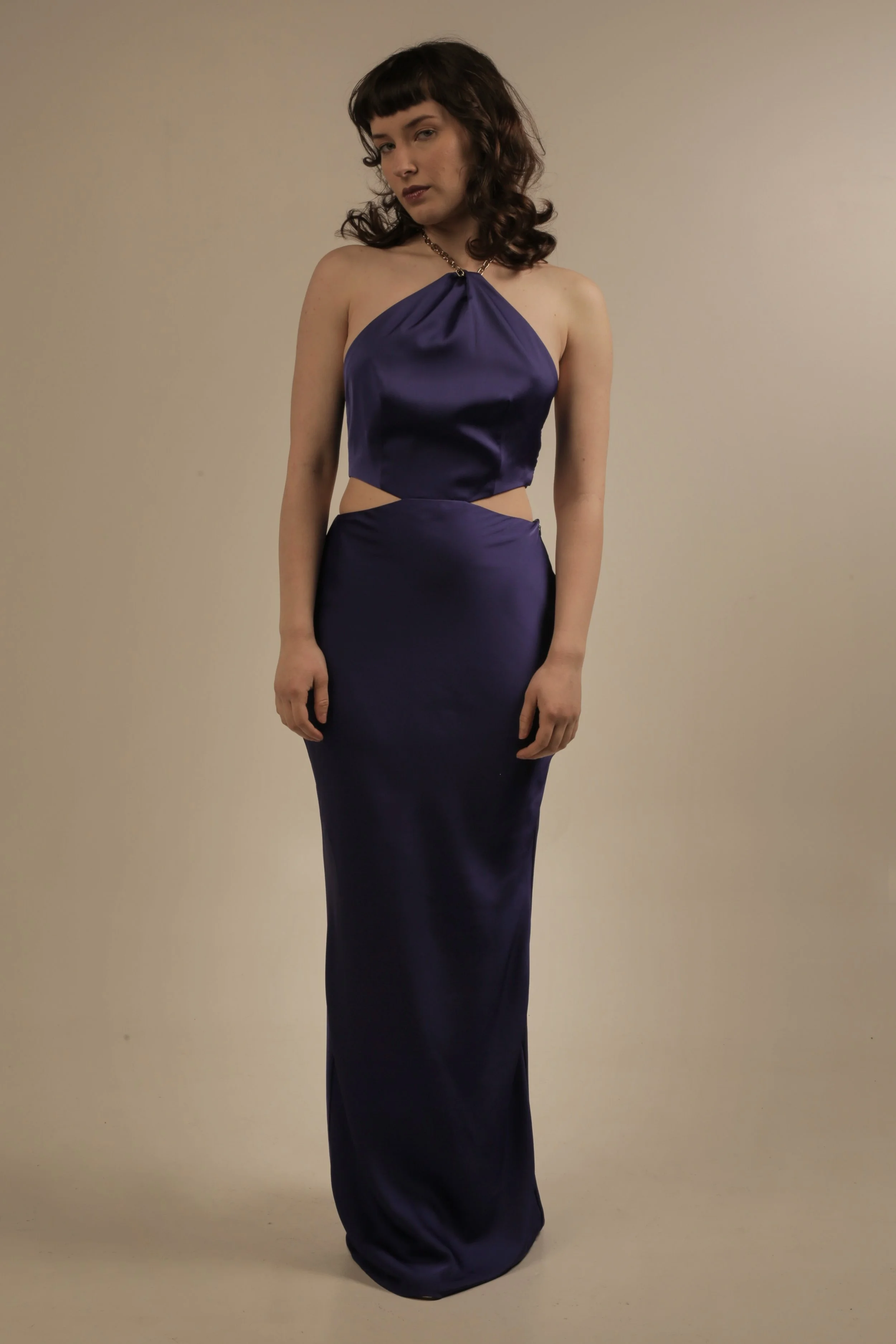 A woman in a dark purple satin evening gown with cutouts at the waist, standing against a plain beige background.