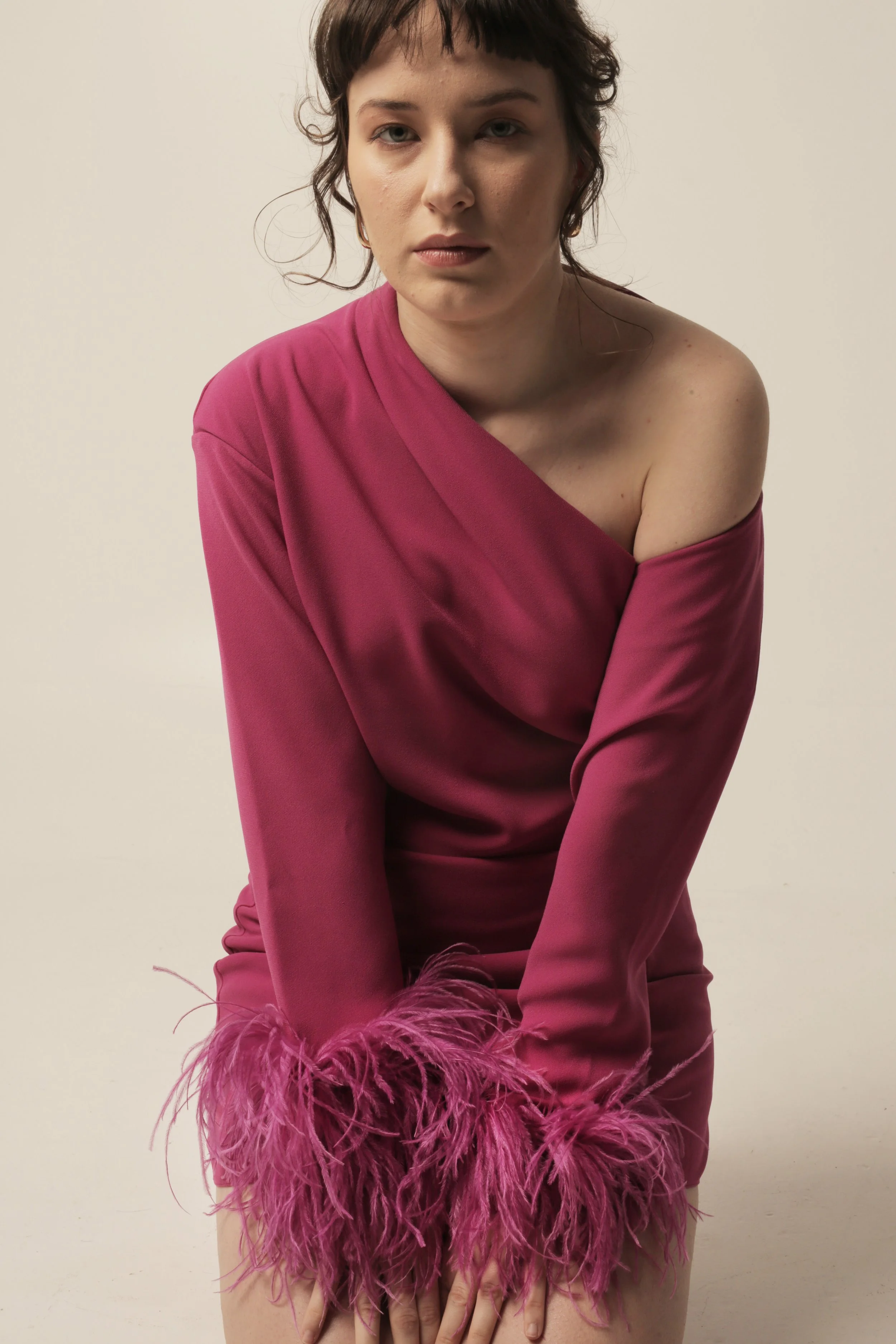 A woman with short wavy dark hair wearing a pink satin dress with feathered cuffs, sitting with her hands on her knees against a plain background.