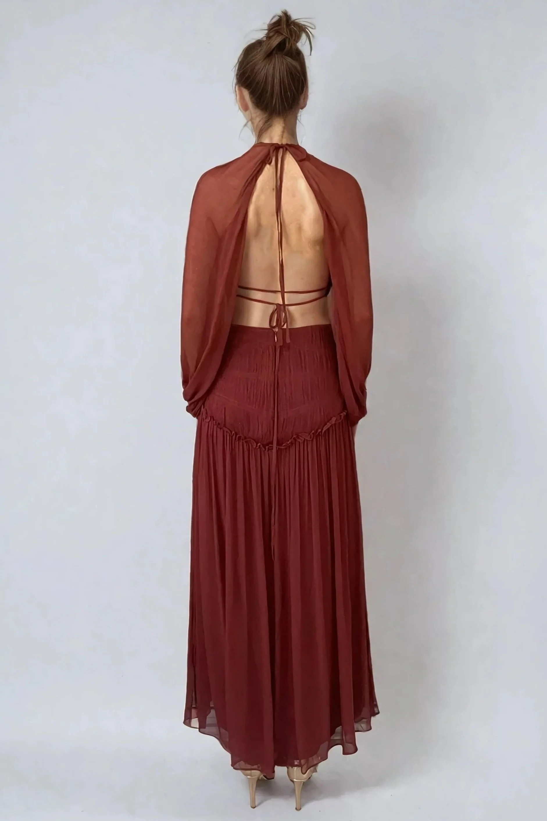 Back view of a woman wearing a sheer rust-colored cape, a strappy bronze top, and a long, pleated rust-colored skirt, standing against a plain wall.