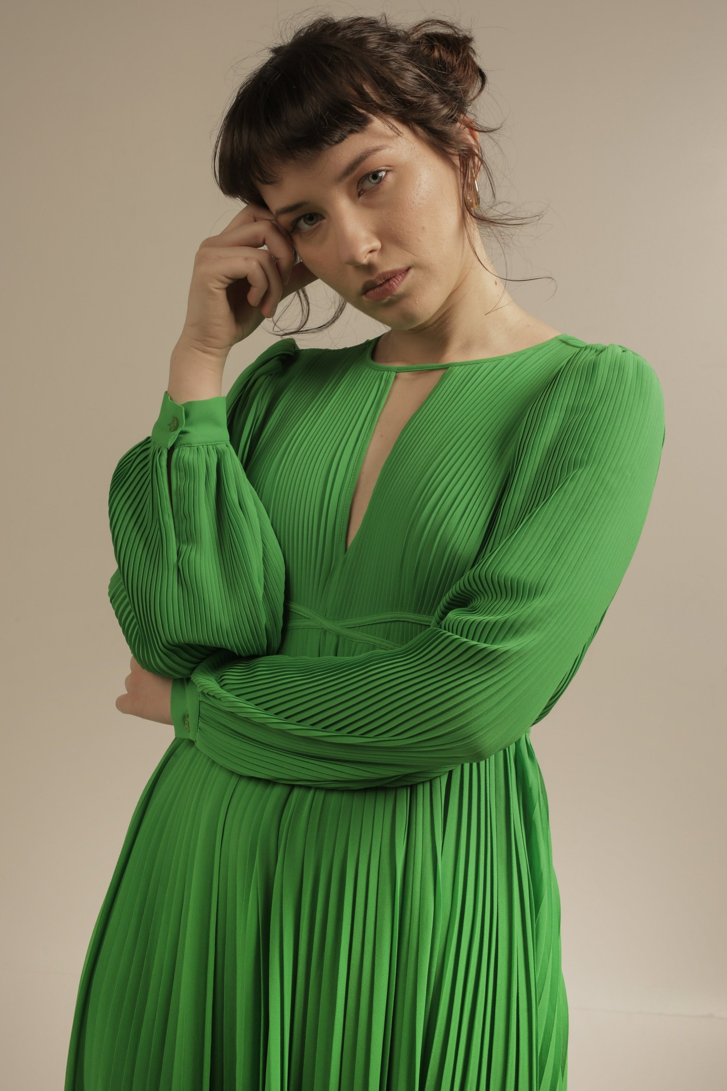 A woman with short brown hair and light skin wearing a bright green pleated dress, standing against a neutral background, looking at the camera with one hand touching her temple.