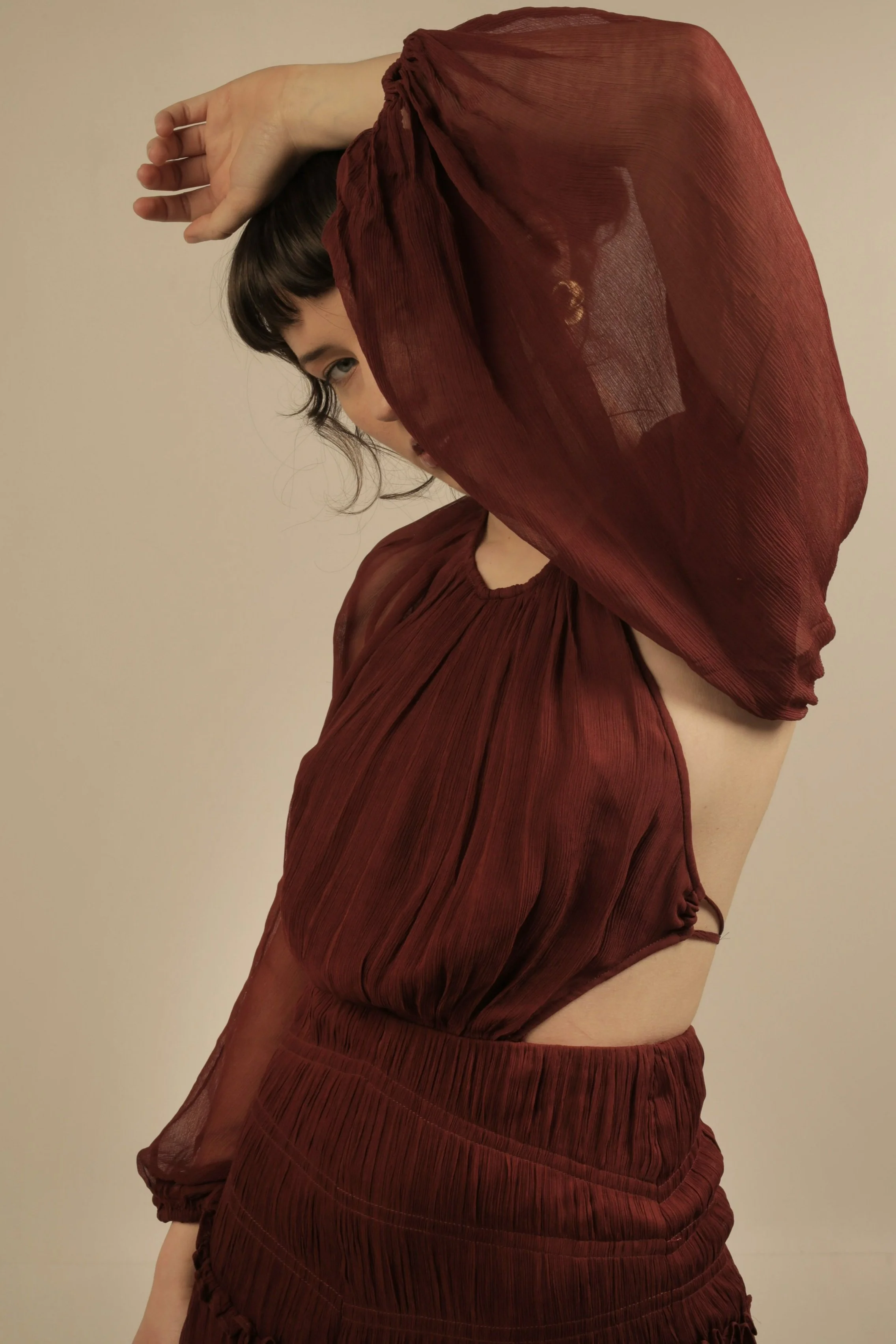 A woman with short dark hair and bangs wearing a sheer, pleated, deep red dress with long sleeves, posing against a plain beige background.
