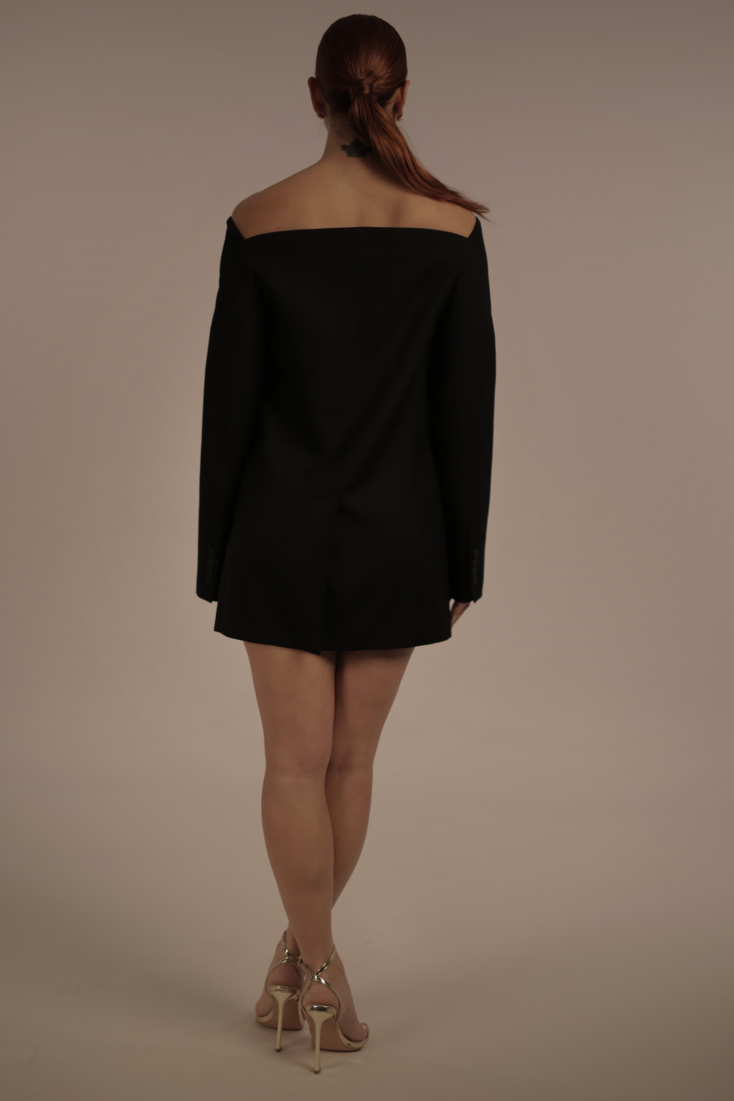 Woman with red hair in a loose ponytail, wearing a black off-the-shoulder blazer with no visible shirt underneath, beige high heels, standing against a neutral background, viewed from behind.