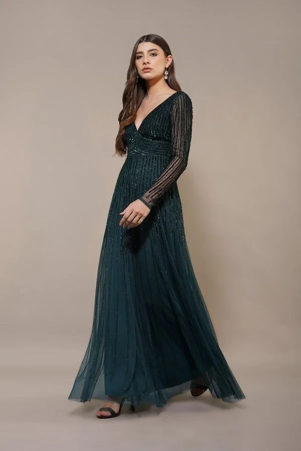 A woman wearing an elegant, long, dark teal evening gown with long, sheer, striped sleeves and embellishments, standing indoors against a plain background.