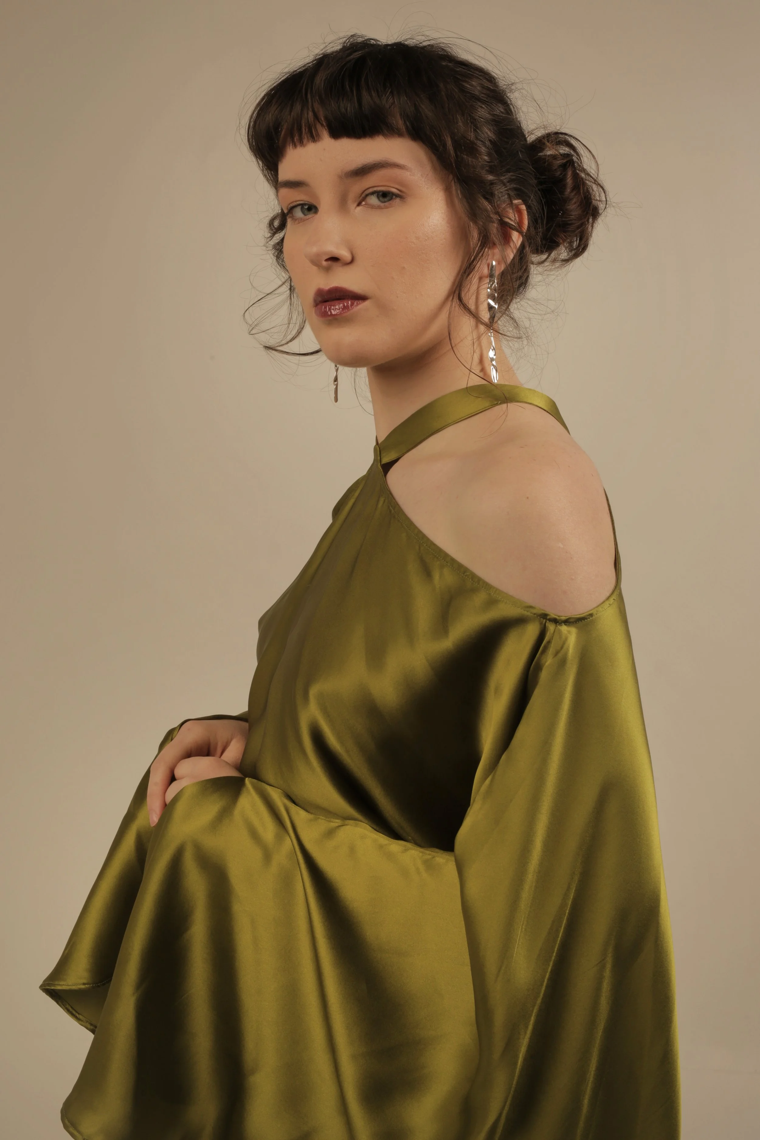 Woman with dark hair styled in a bun, wearing an olive green satin dress with open shoulders and dangling earrings, against a plain background.