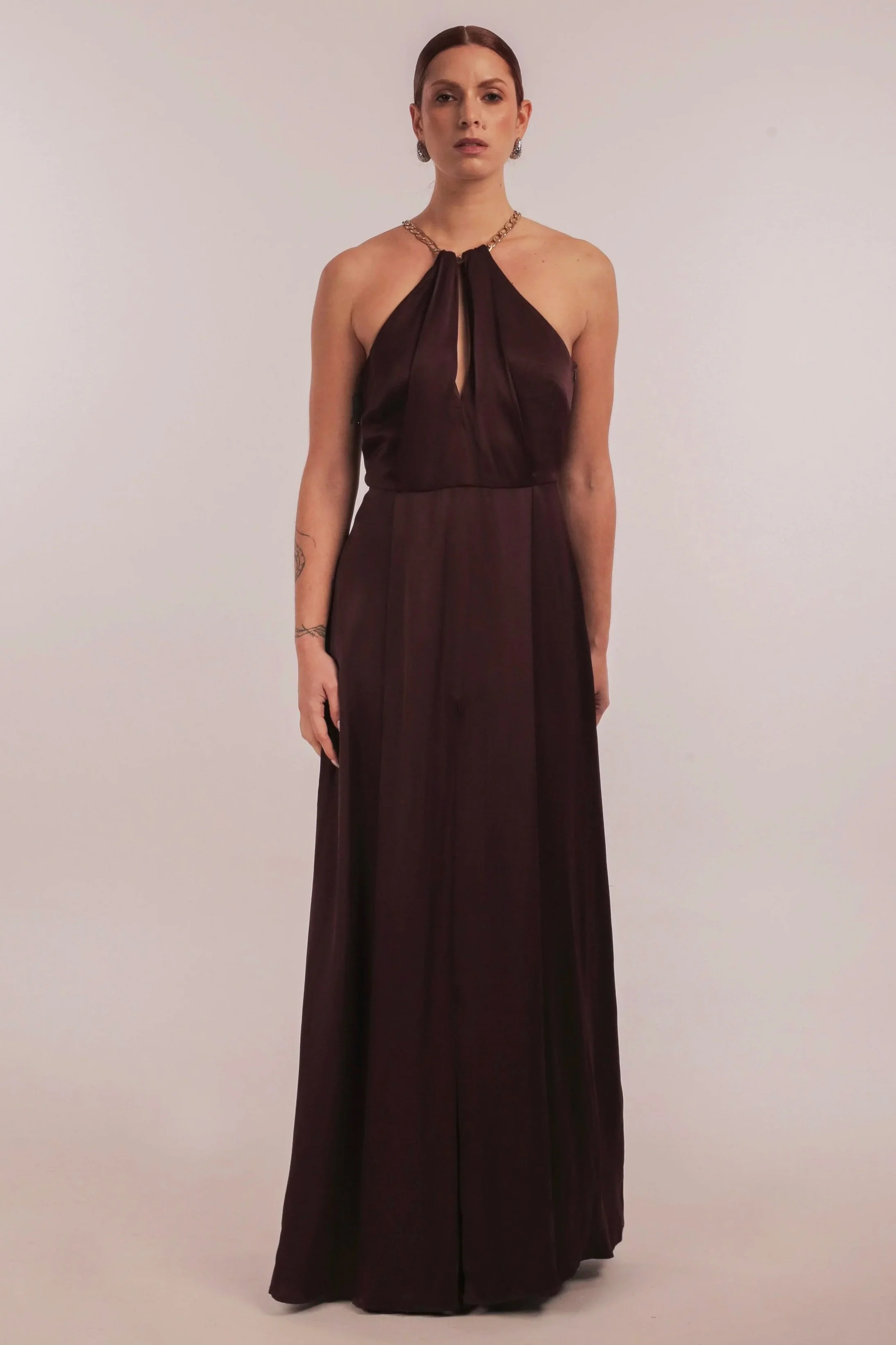 Woman in a dark mauve satin halter neck evening gown with gold jewelry standing against a plain background.