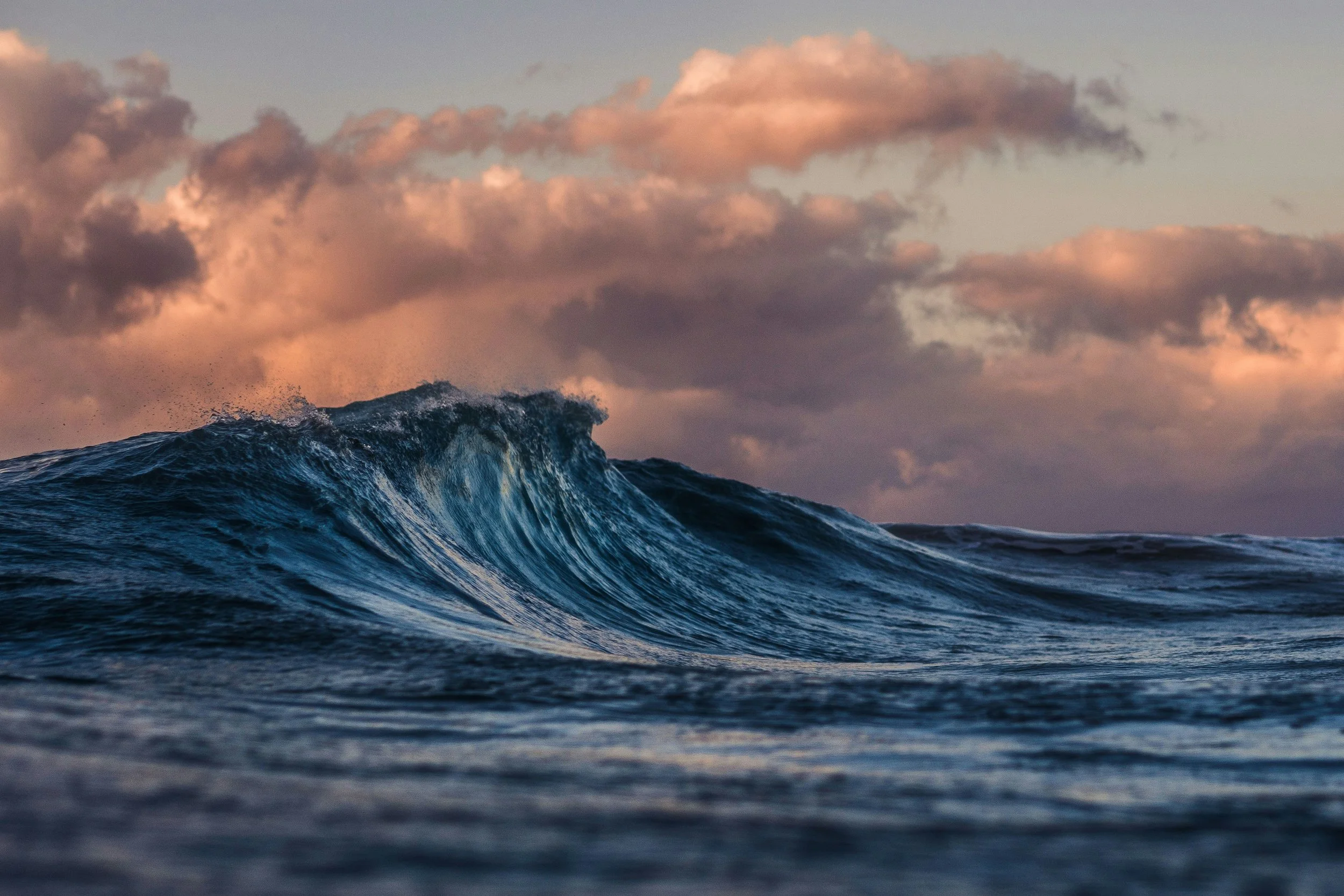 Fotografie van een oceaangolf met een kleurrijke wolkenlucht in de achtergrond, waarschijnlijk tijdens zonsopgang of zonsondergang.