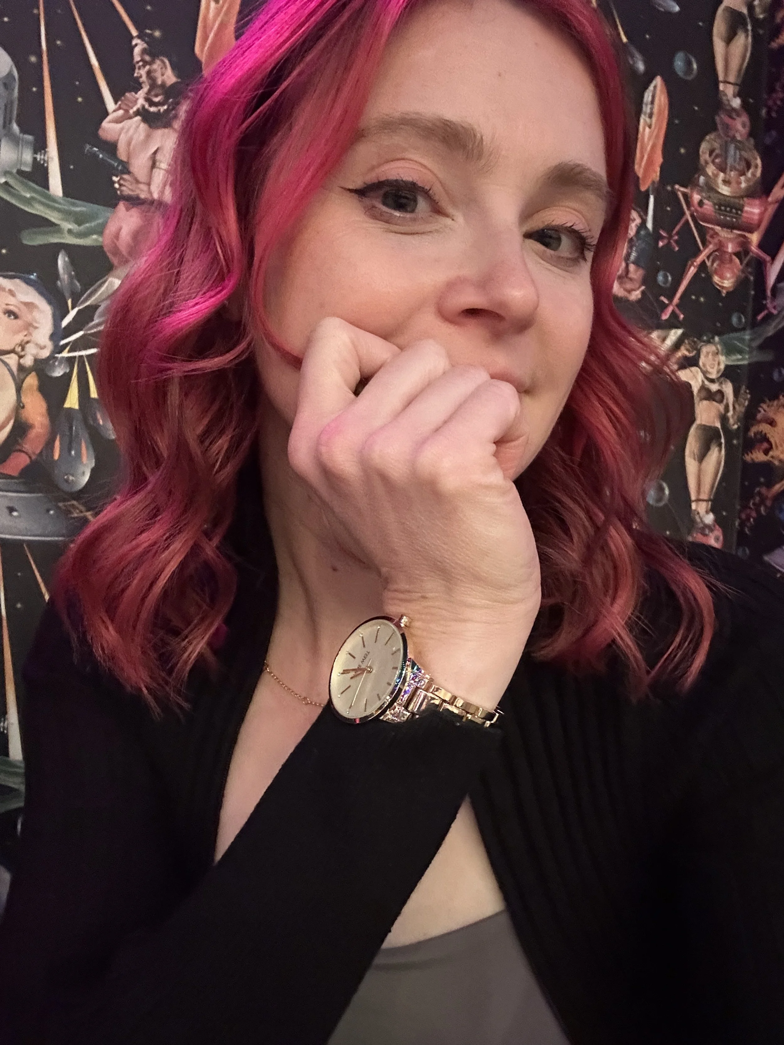 A woman with pink dyed wavy hair, wearing a gold wristwatch, a delicate gold necklace, and a dark sweater, resting her face on her hand, in front of a black wall with colorful, vintage-style posters and artwork.