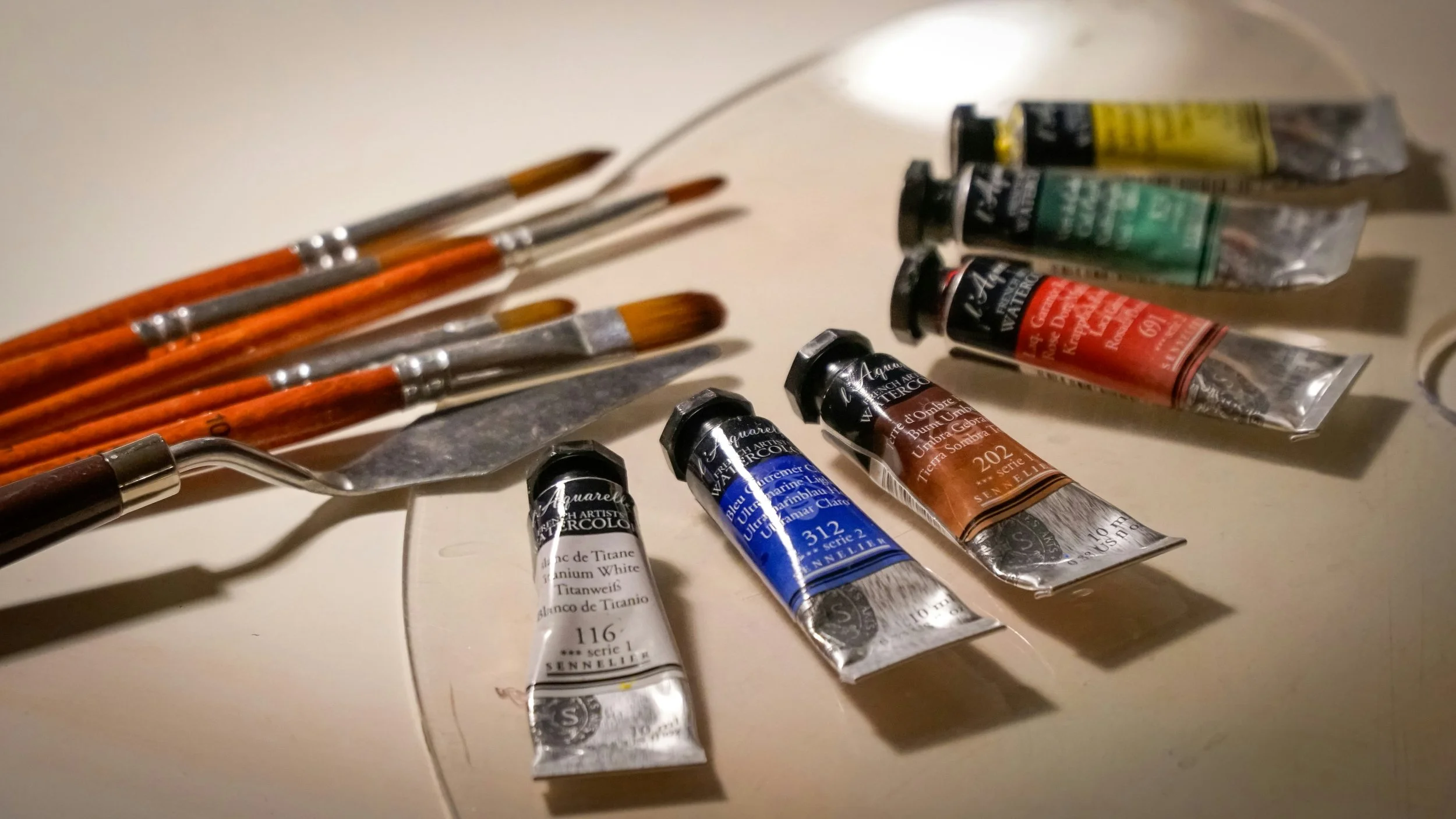 Set of watercolor paint tubes in various colors and paintbrushes arranged on a light-colored surface.