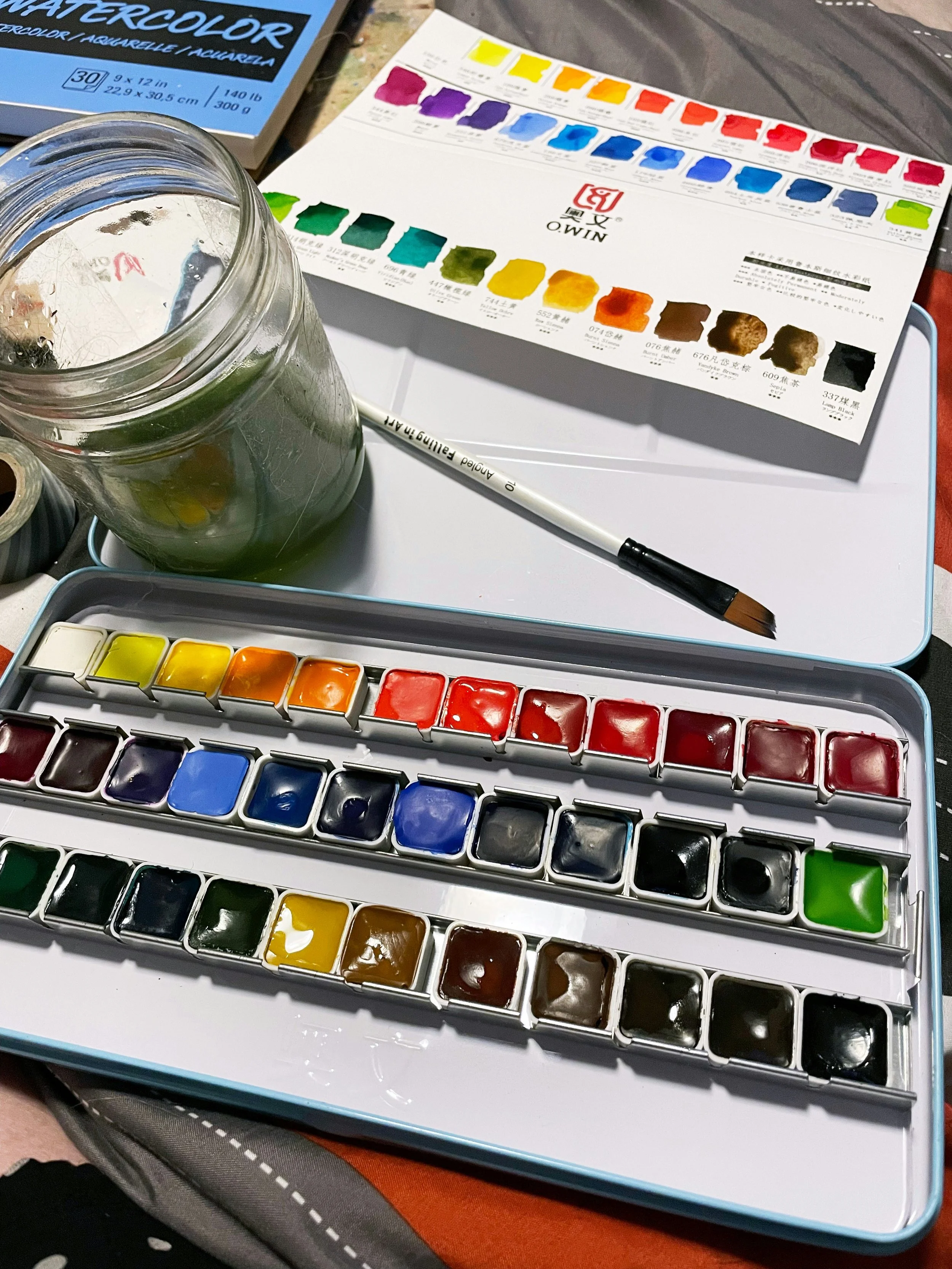 A watercolor paint set with various colors, a color chart, a paintbrush, a glass jar with water and paint, and art supplies on a table.