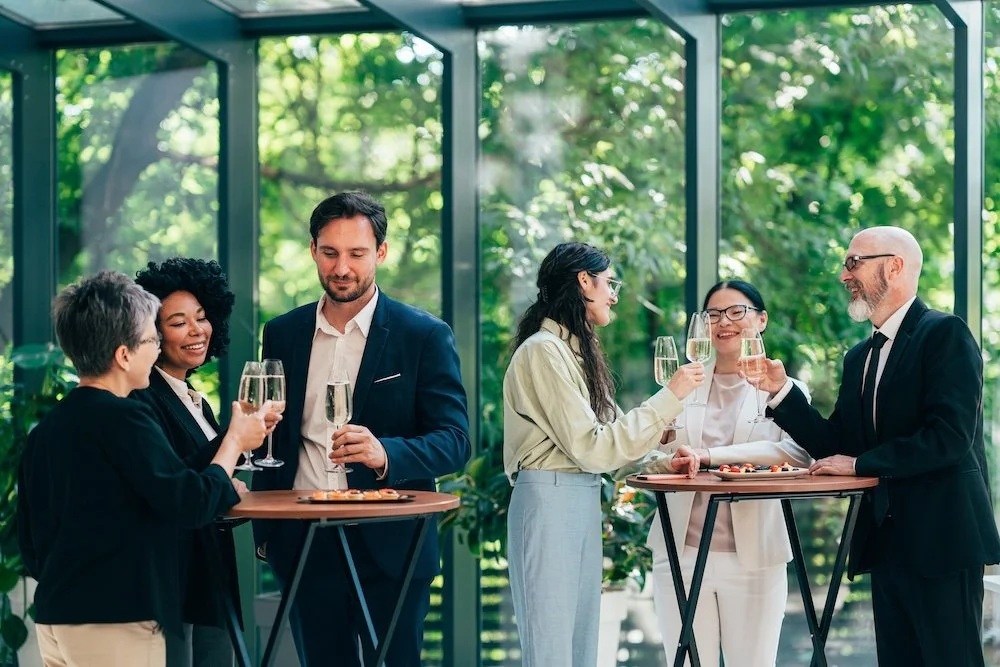 Group of co-workers socializing and toasting in a bright room with greenery outside.