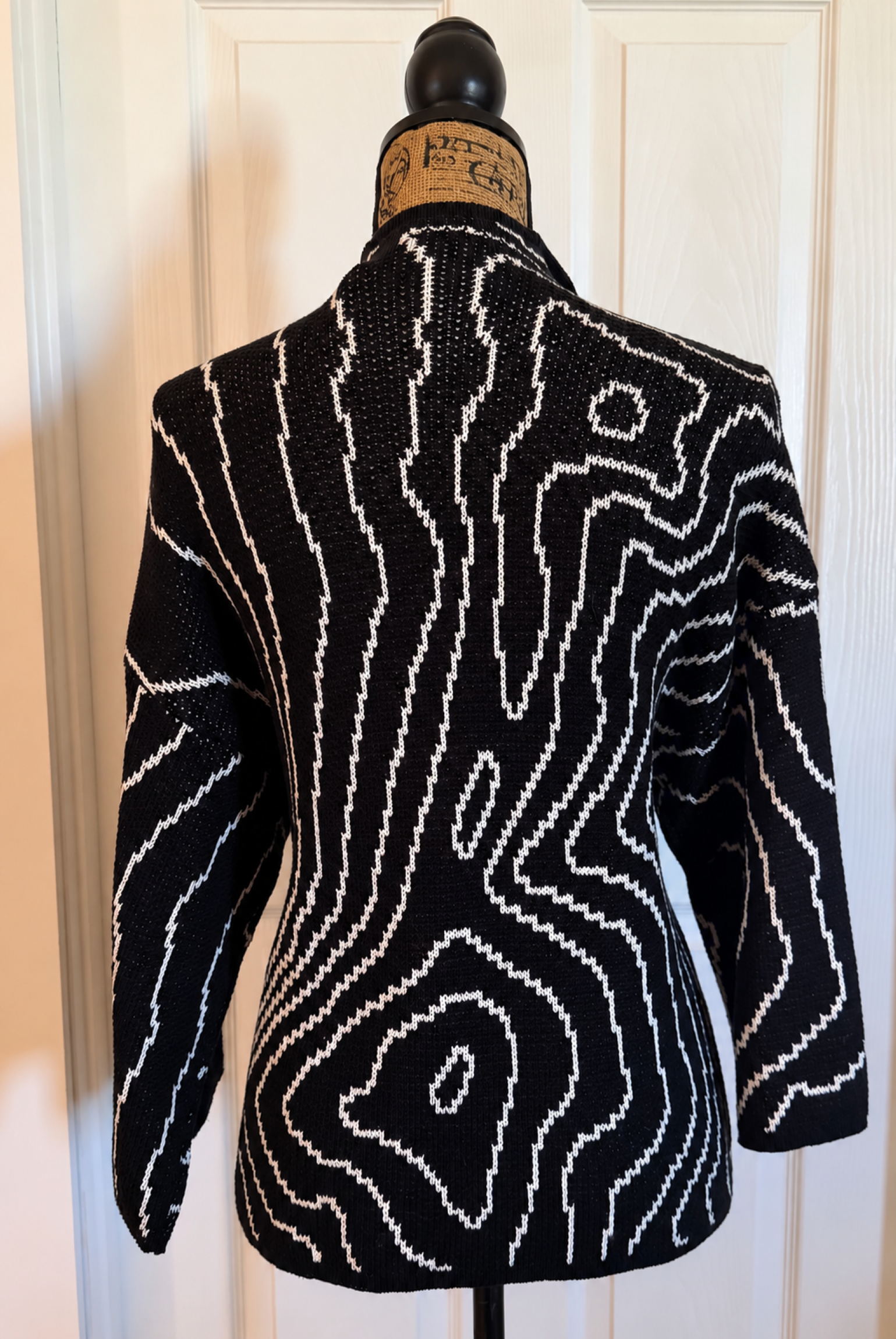 Back view of The Chickadee Tree Maroon Bells Cartography knit jacket featuring white topographic contour lines against black fabric, depicting the landscape of the Maroon Bells in Colorado.