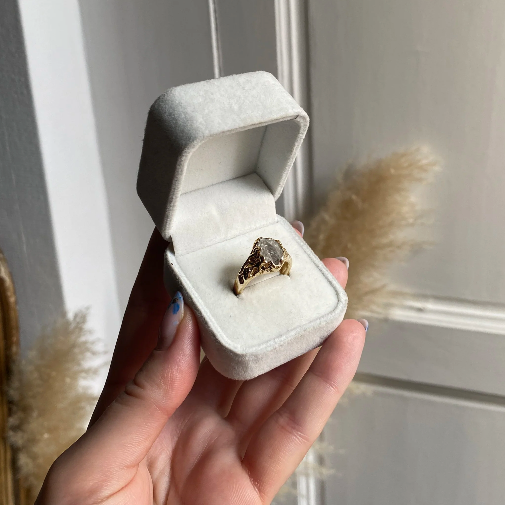 Une main tient une boîte à bijoux en velours blanc contenant une bague en or avec une pierre précieuse. En arrière-plan, une porte et des éléments décoratifs feuillus.