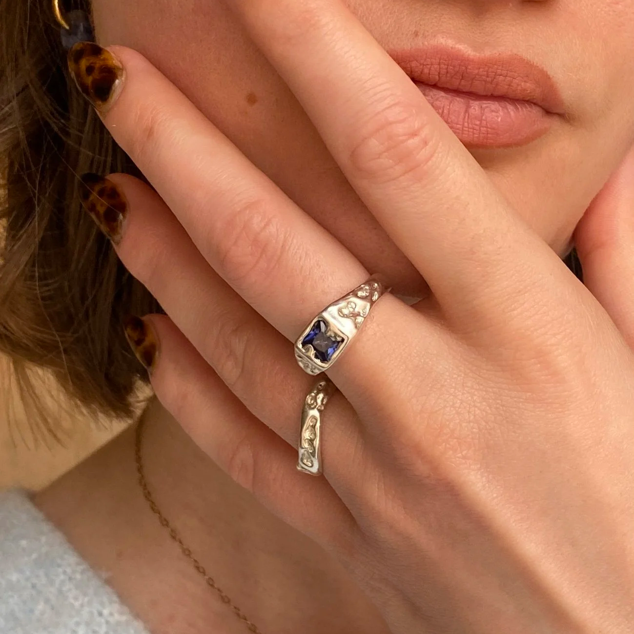 Une femme avec des ongles à motif tacheté et portant des bijoux, dont deux bagues en argent avec des pierres précieuses et une chaîne dorée avec pendentif solaire, près de son visage et de ses lèvres