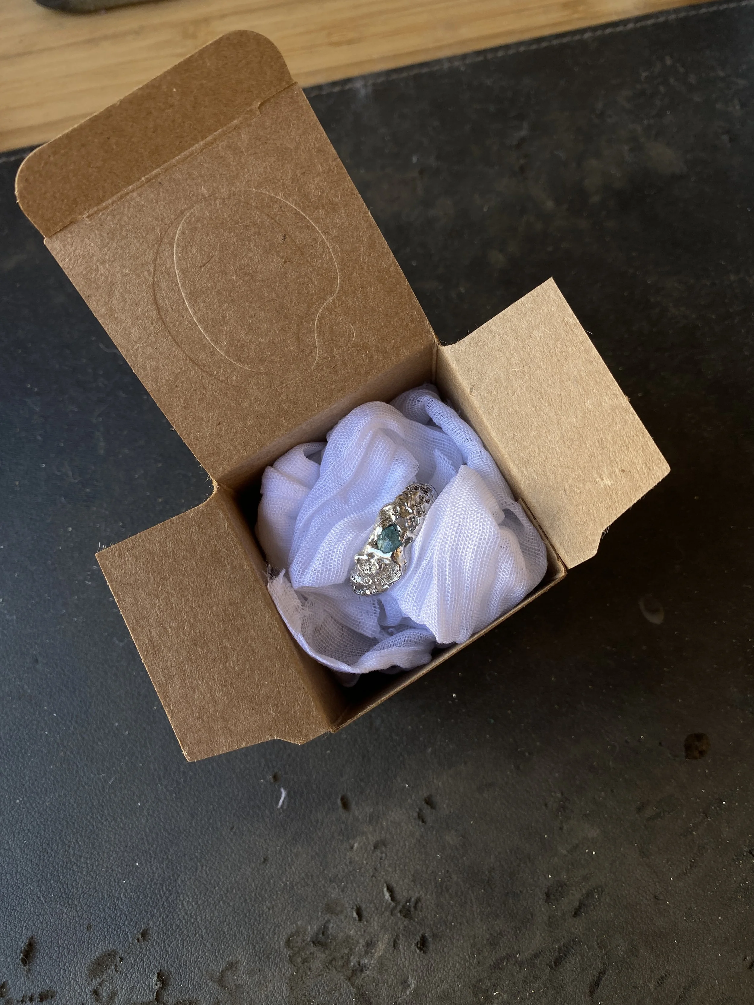 Une boîte en carton contenant une bague en argent ornée de pierres, entourée de tissu blanc, posée sur une surface sombre.