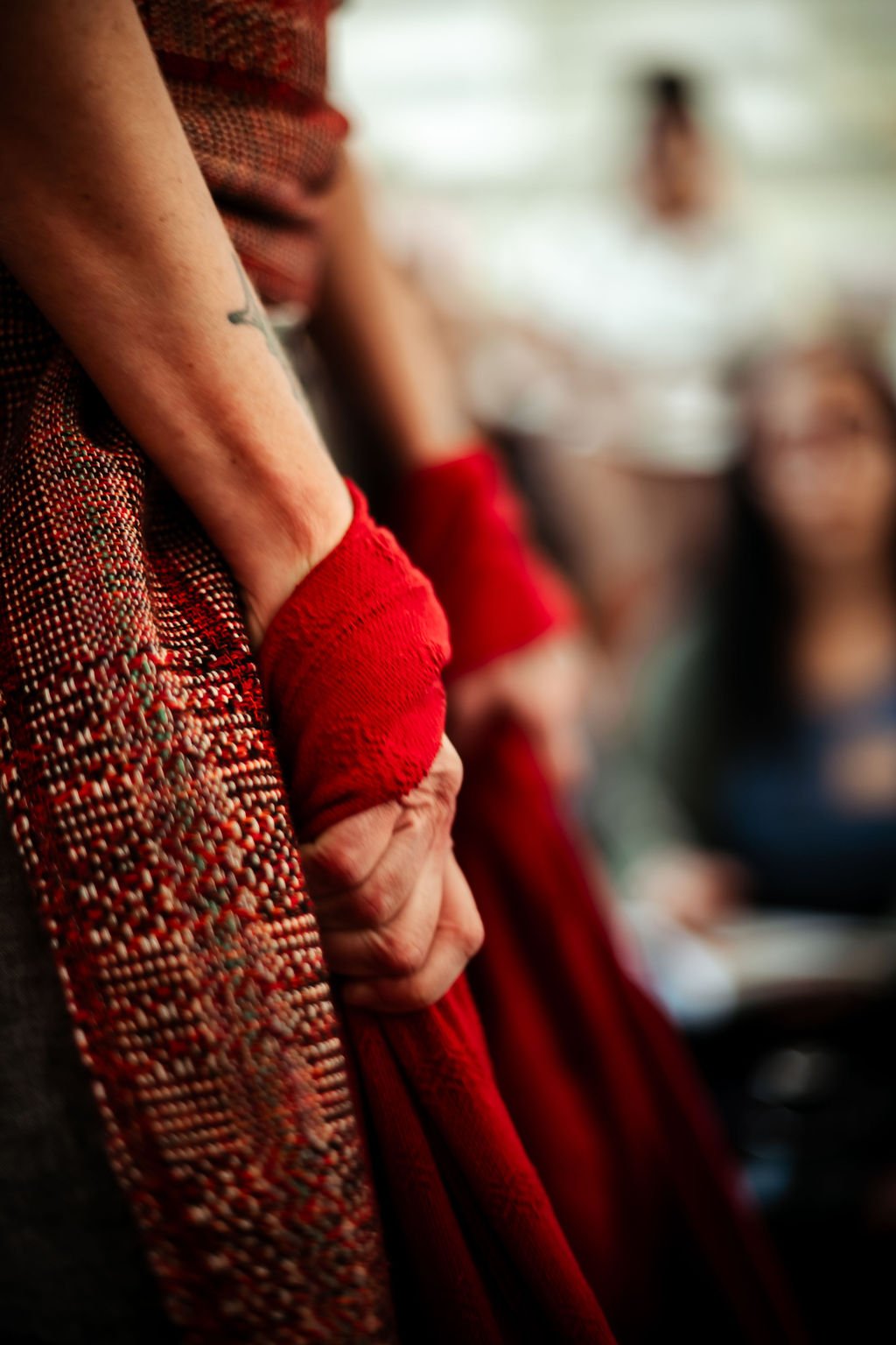Close-up of a person's hand with a red rebozo, standing against a blurred background of other people in a closing of the bones ceremony setting.