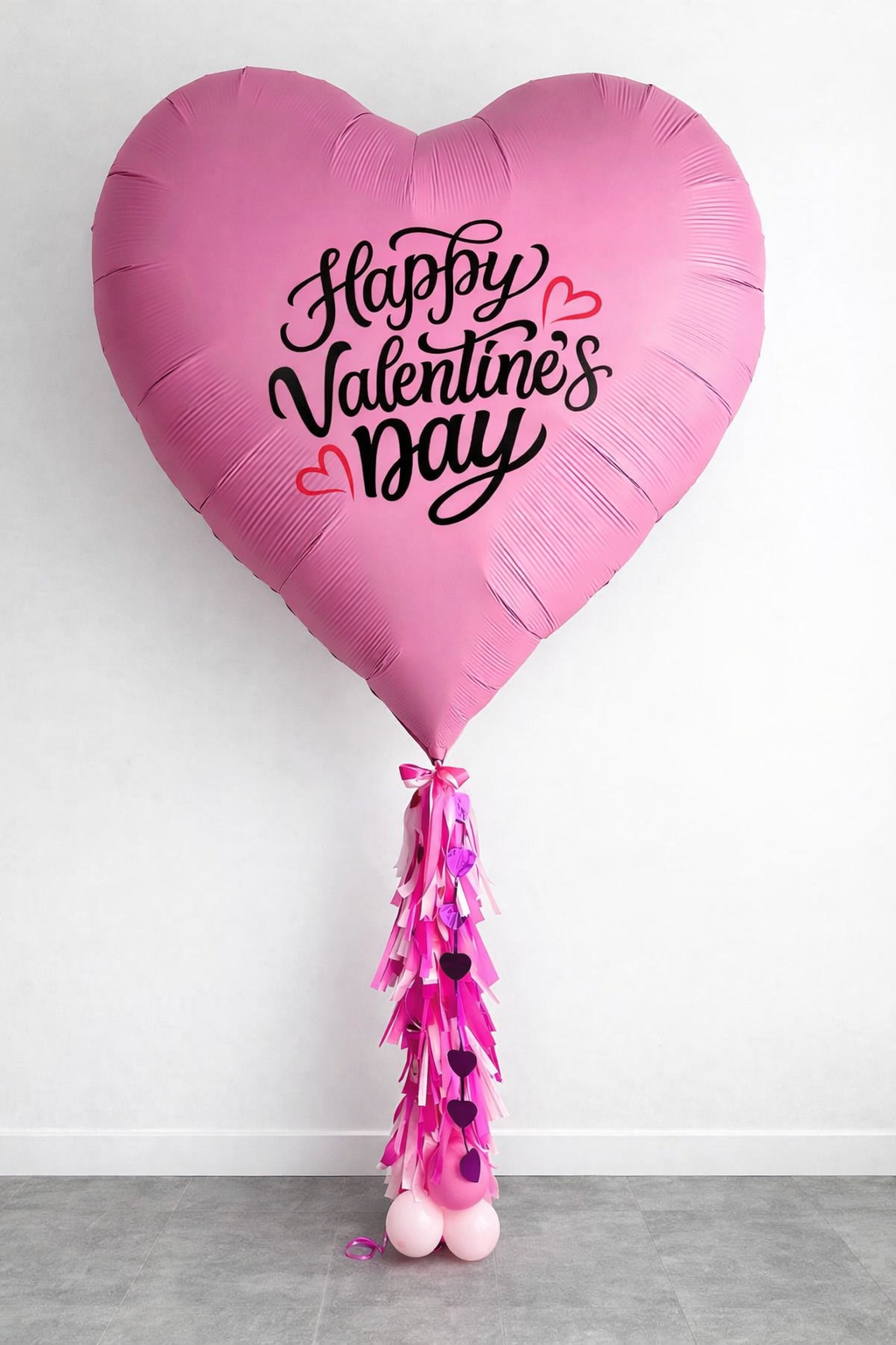 Pink heart-shaped balloon with 'Happy Valentine's Day' written in black cursive and small red and pink hearts, attached to a pink and purple tassel made of paper and foil, with pink and white balloons at the base.