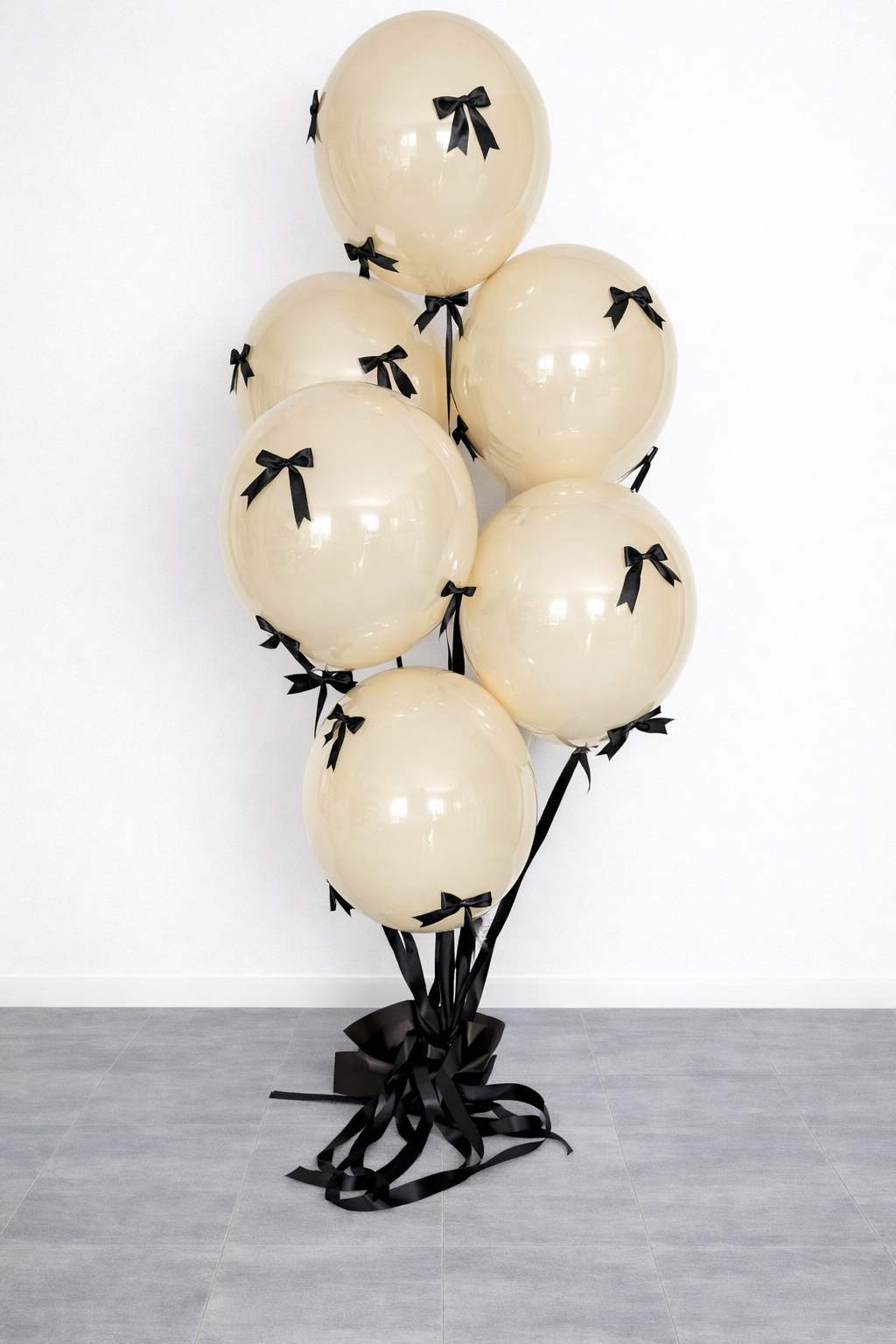 A bundle of cream-colored balloons decorated with small black bows tied around each balloon, tied together with black streamers, and placed against a plain white wall.