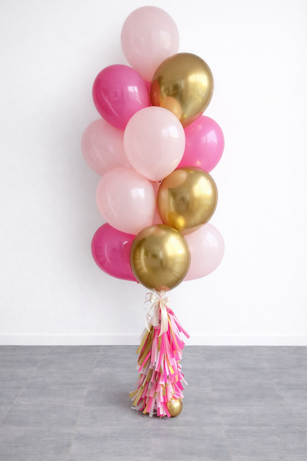 A bouquet of pink and gold balloons tied with pink and beige ribbons, with a tassel made of pink, beige, and gold ribbons at the bottom, placed on a gray tiled floor against a white wall.