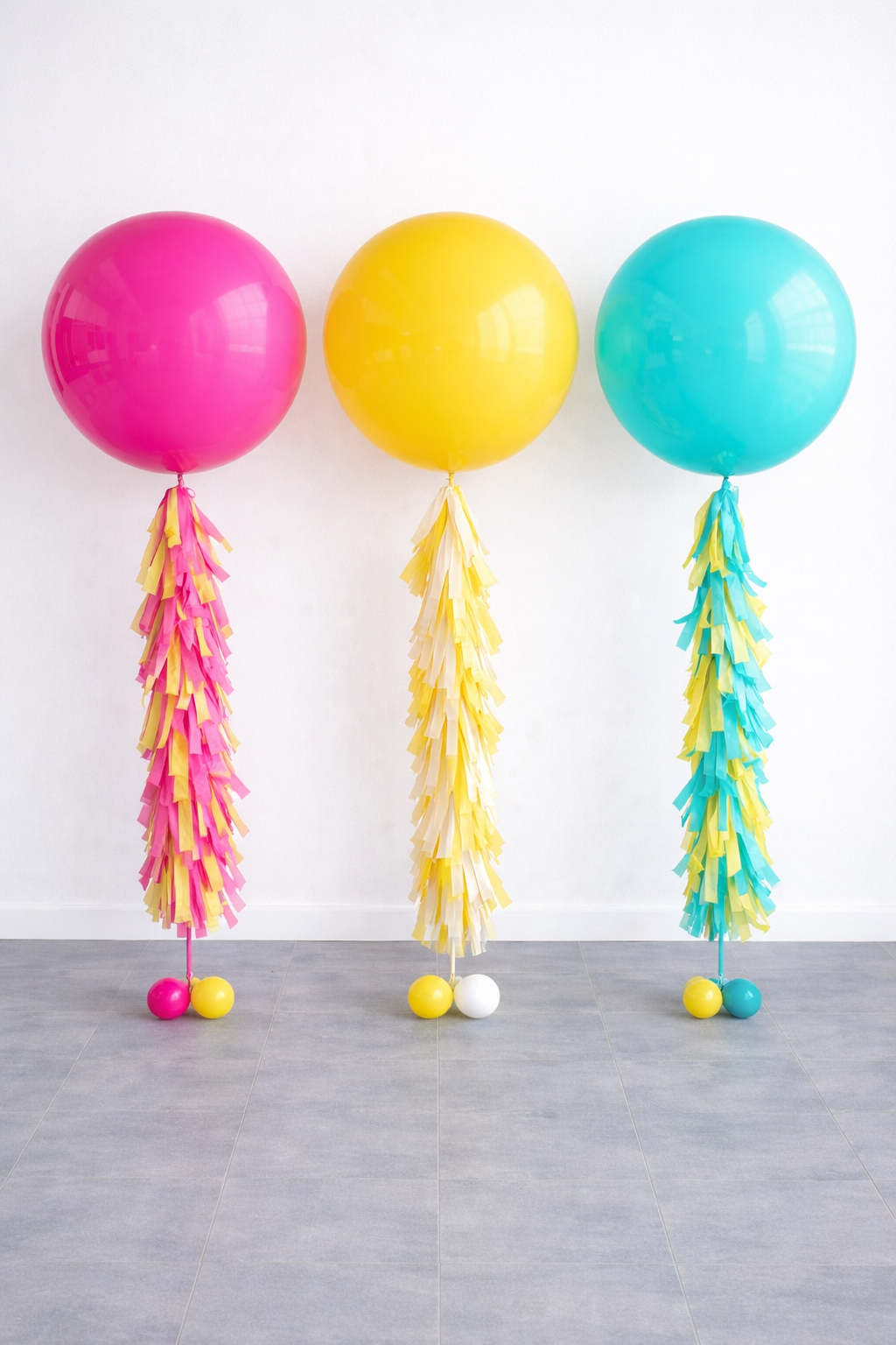 Three large colorful balloons (pink, yellow, blue) each with a tassel tail and two smaller balloons at the base, positioned against a plain white wall.