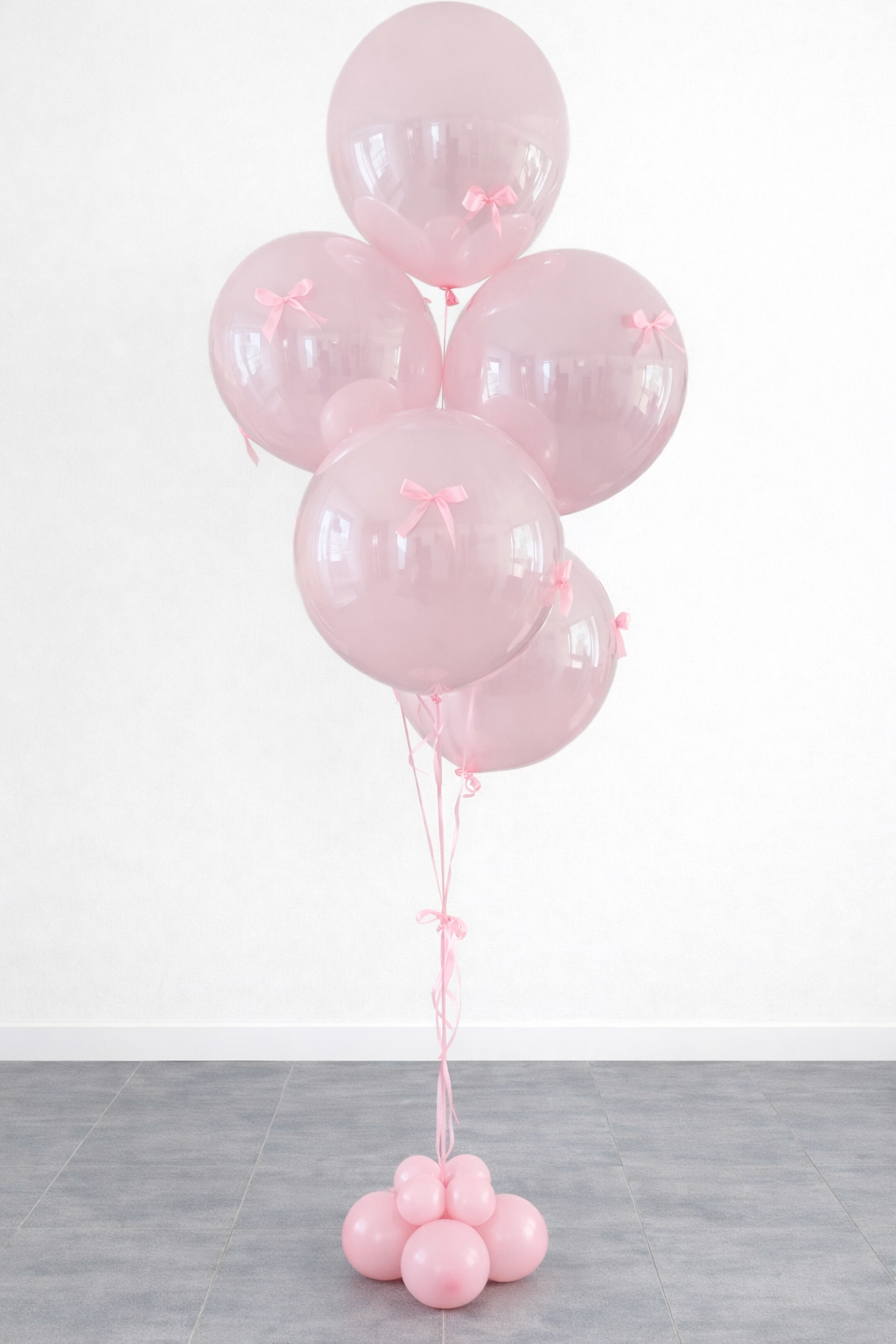 Pink balloons with small bows tied to each balloon, tied together with pink ribbons, and resting on a cluster of smaller pink balloons on the floor.
