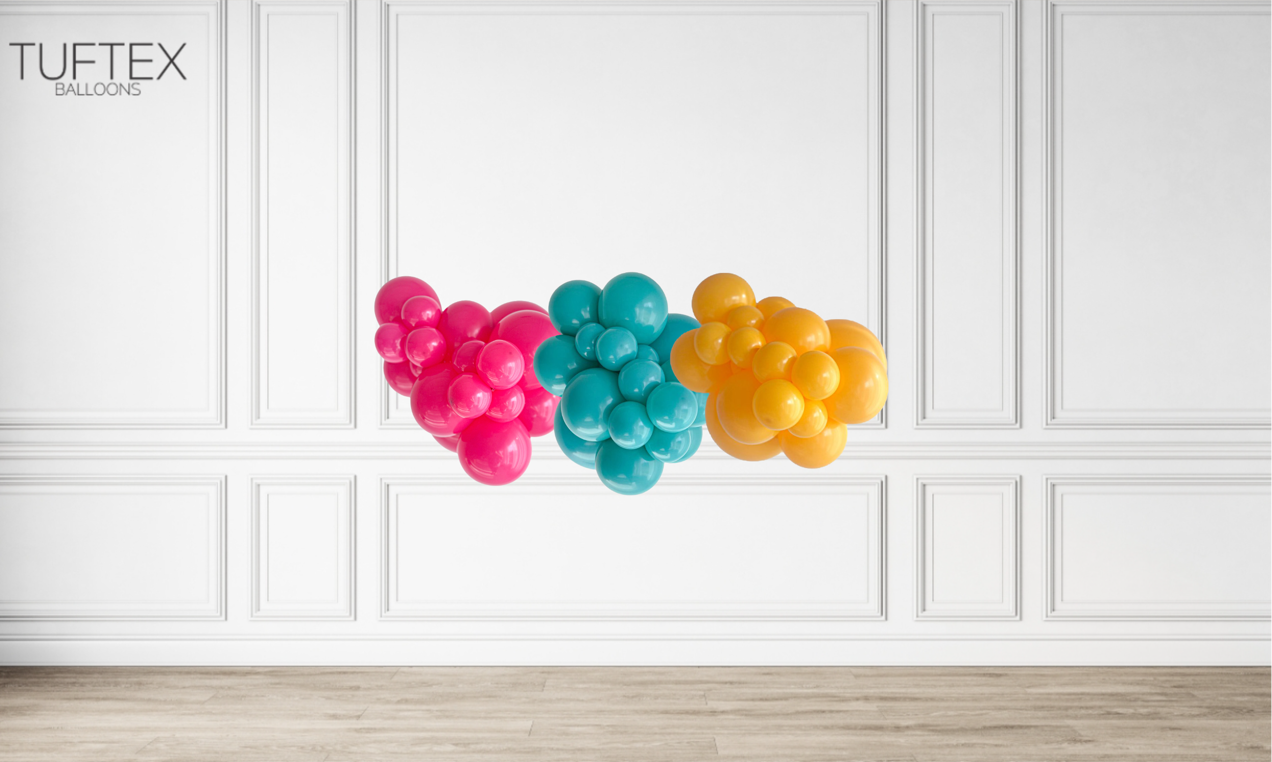 Three clusters of colorful balloons, pink on the left, blue in the middle, and orange on the right, floating against a white paneled wall with light wooden floor.