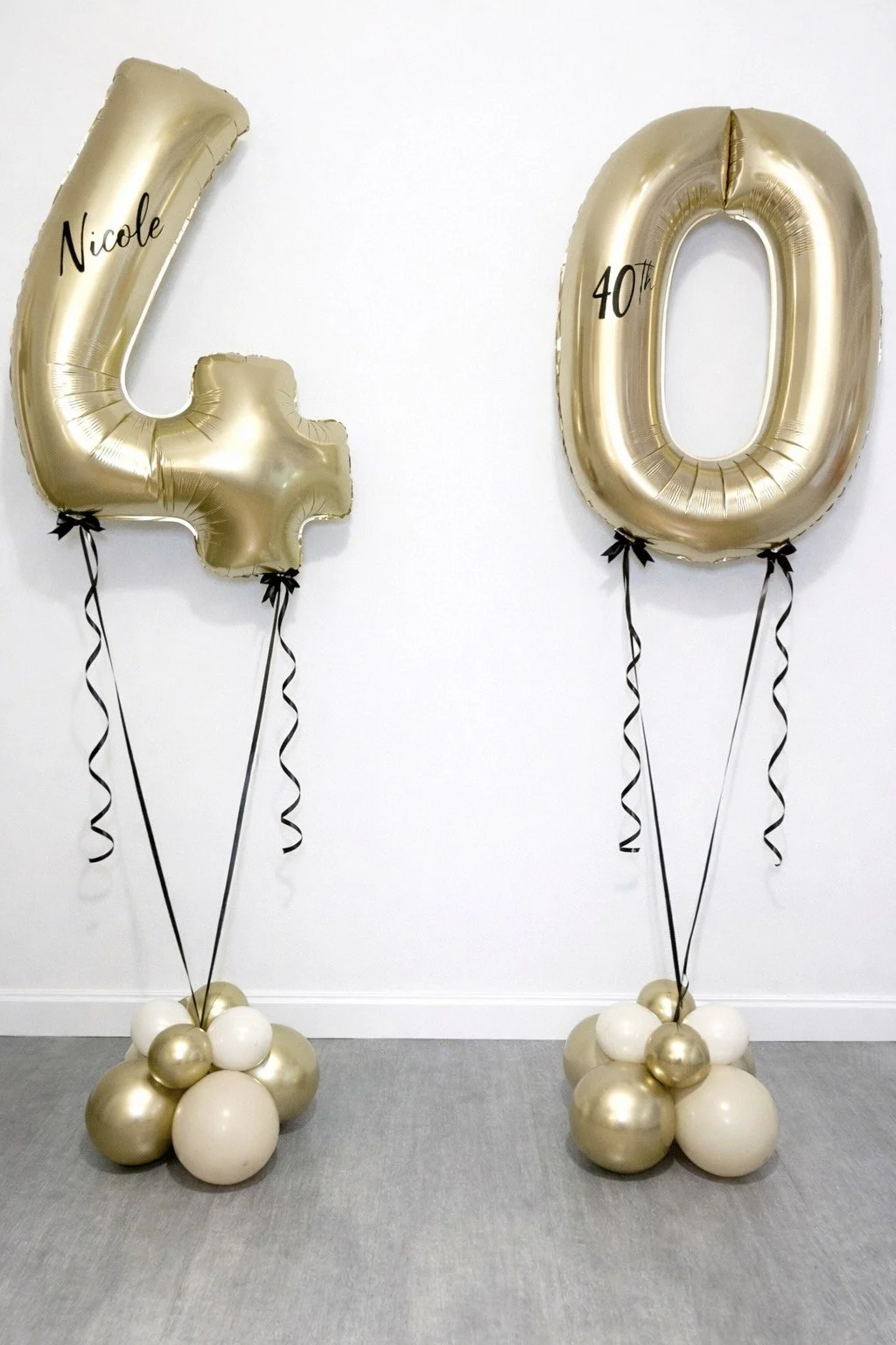 Gold and white balloon decorations shaped as the number 40, with the name 'Nicole' on the left balloon, and balloons anchored with clusters of smaller gold and white balloons at the base, set against a white wall and grey carpet.