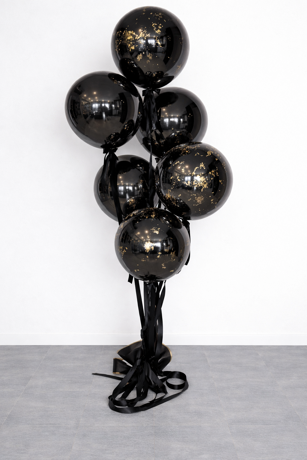 Black balloons with gold speckles tied together with black ribbons and arranged in a bouquet, standing against a plain white wall.
