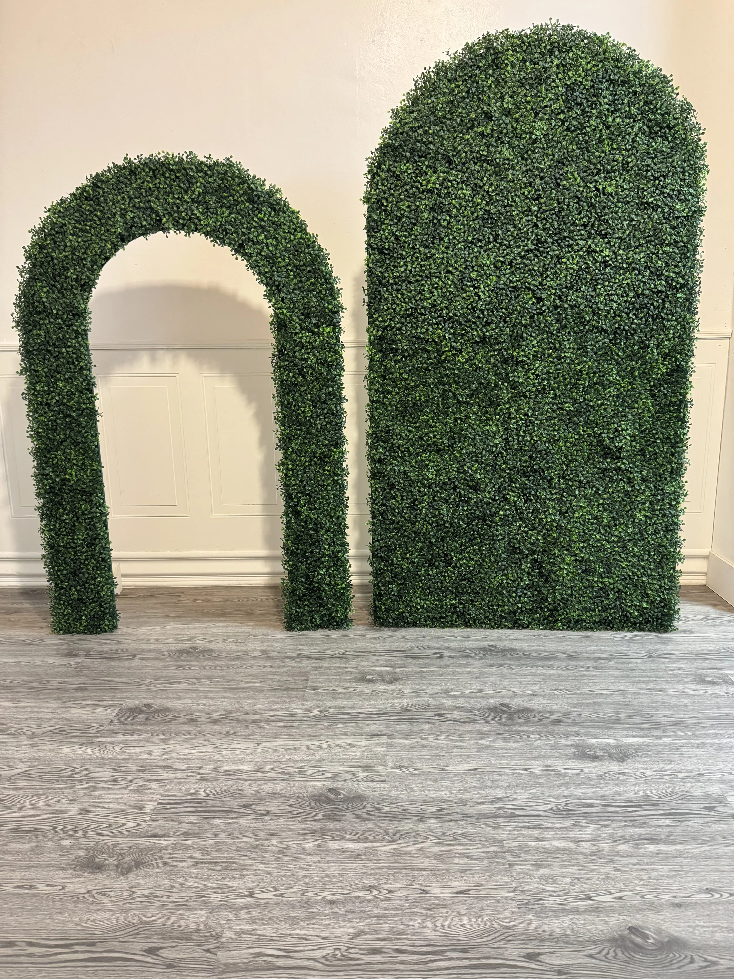 Green artificial hedge structures arranged in an arch and a rectangular shape, placed indoors against a white wall with grey wood flooring.