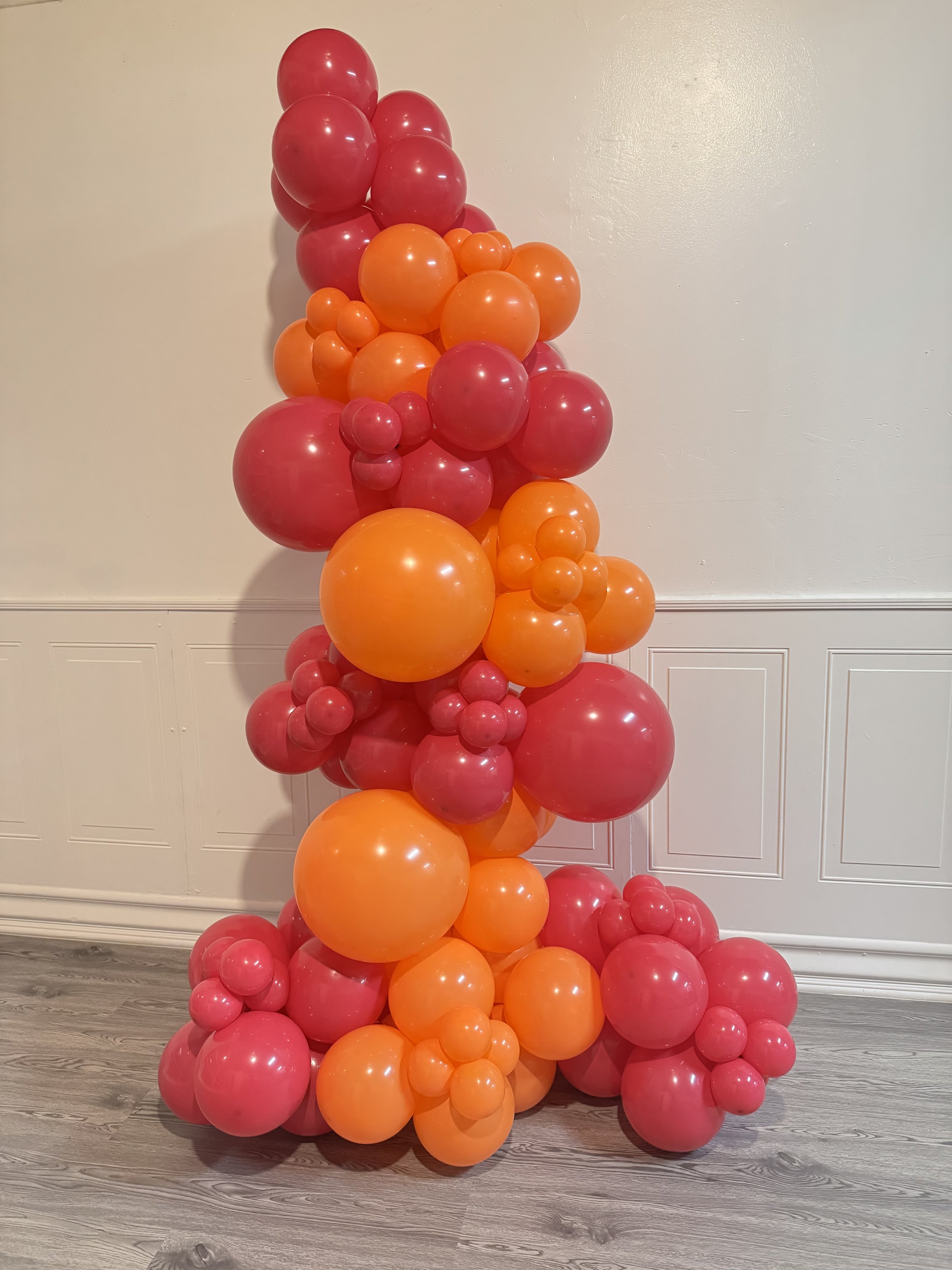 A colorful balloon sculpture with a mix of pink and orange balloons of various sizes stacked against a white wall with white wainscoting and a gray wood floor.