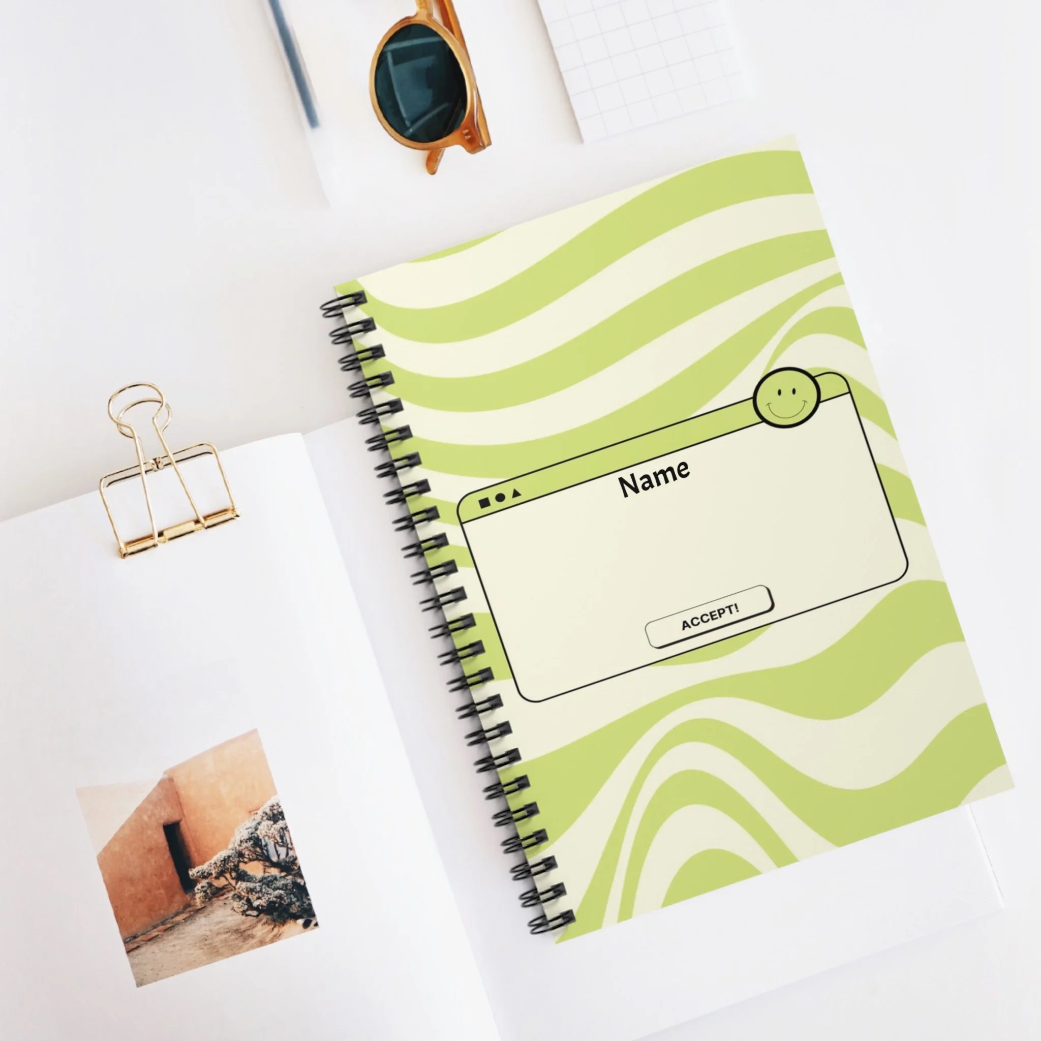 A spiral-bound notebook with a green and white wavy pattern cover, featuring a mock computer window with a smiley face, a space for writing a name, and an accept button. The notebook is on a white desk next to a photo, a gold paper clip, a pair of sunglasses, a pen, and a graph paper notepad.