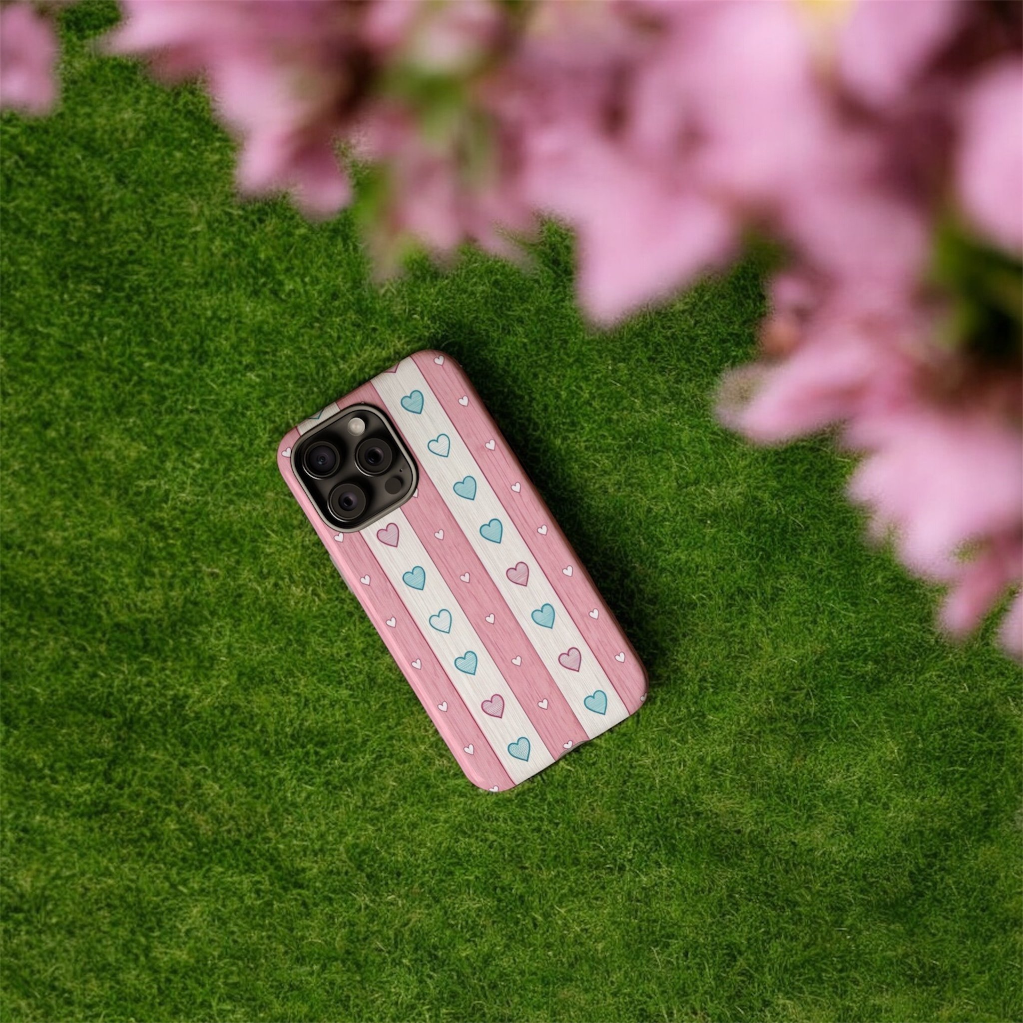 iPhone with a pink case decorated with pink, blue, and white hearts, lying on green grass with pink flowers in the foreground.