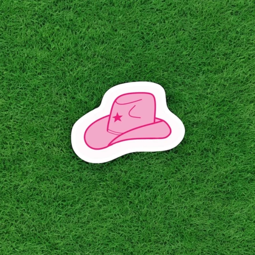 Pink sticker of a cowboy hat with a star and a mustache, placed on green grass.