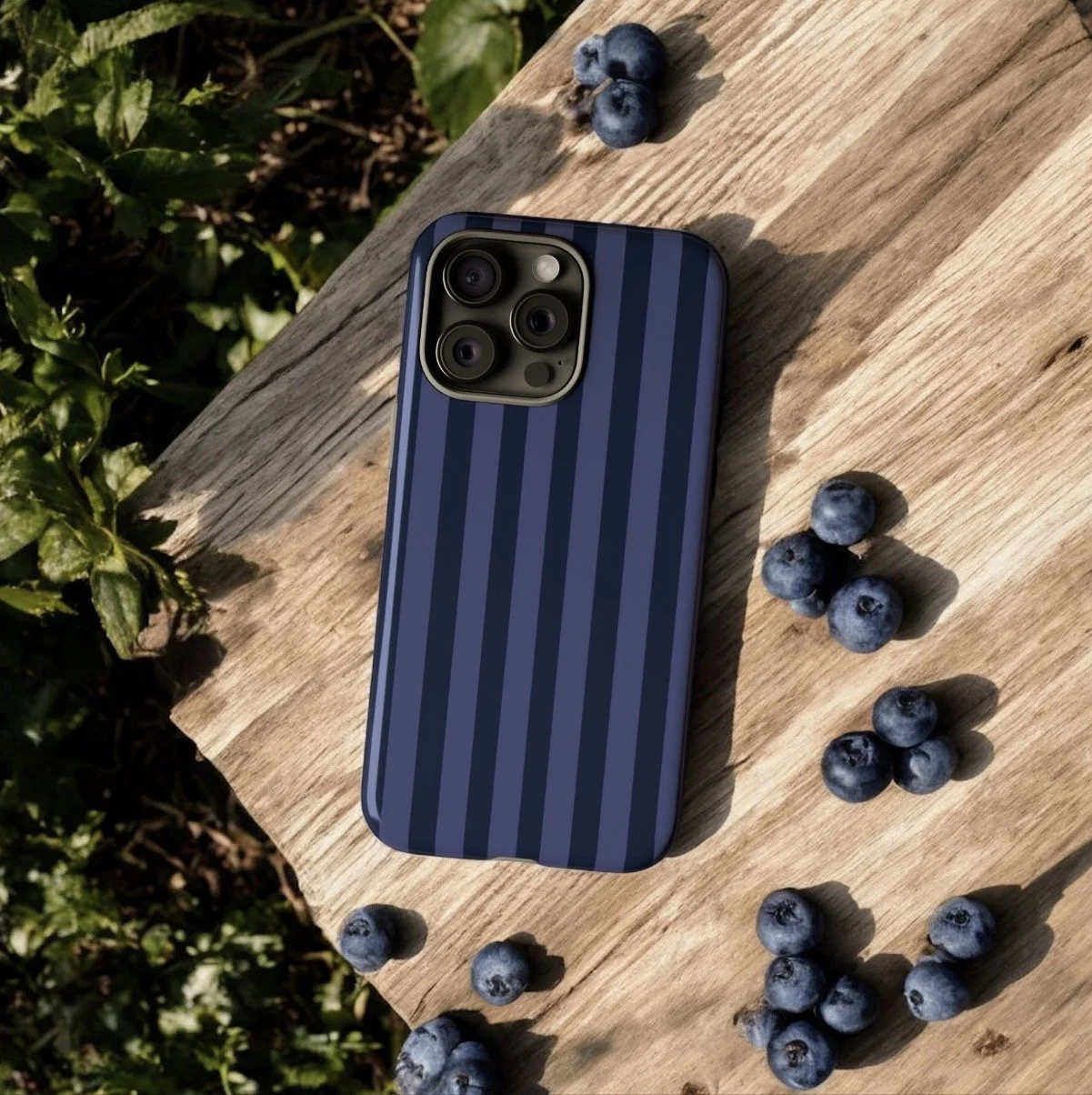 A smartphone with a blue and black striped case is placed on a wooden surface surrounded by scattered blueberries. Sunlight casts shadows, and green foliage is visible in the background.
