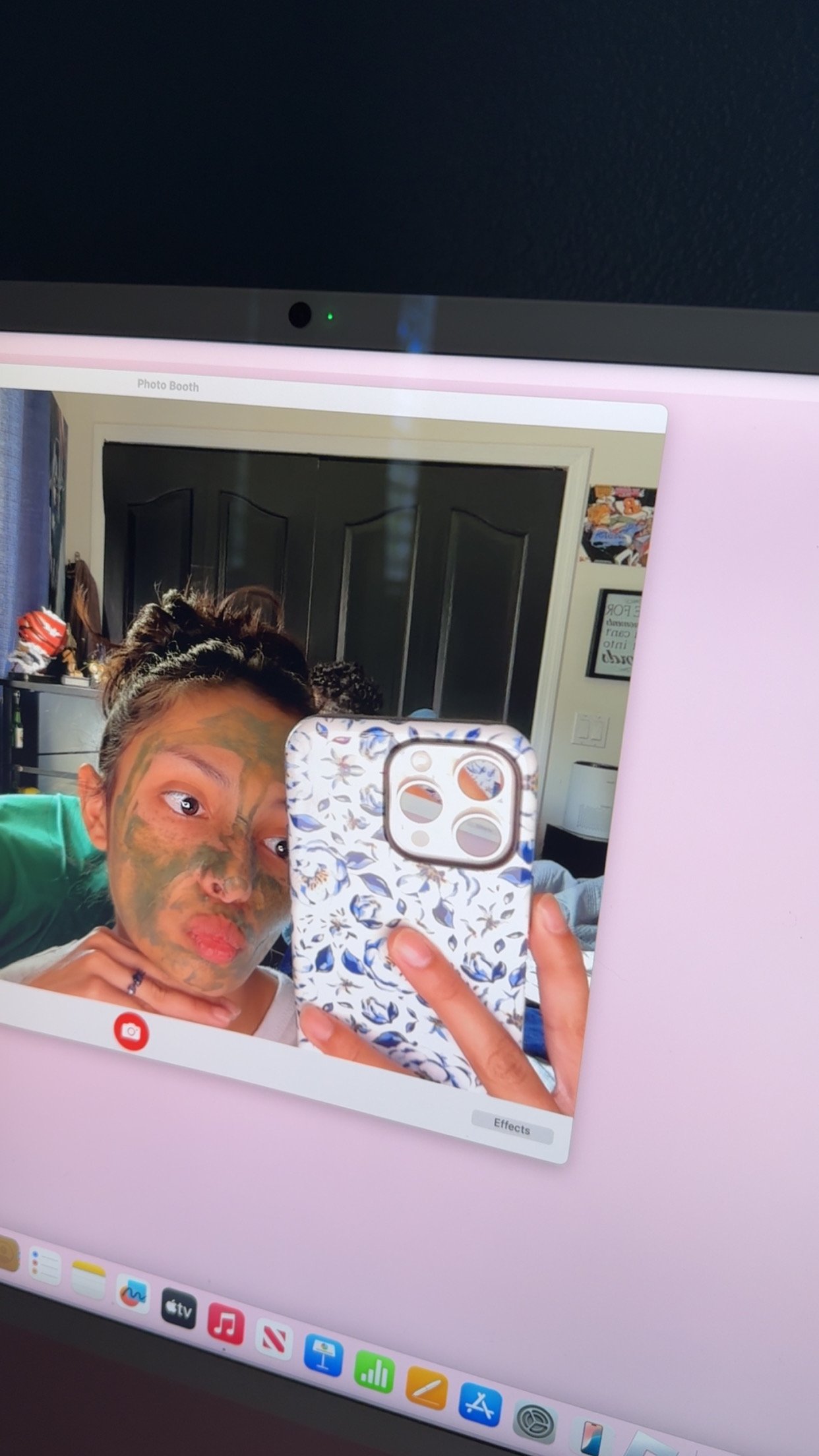 A person taking a selfie with a child who has green face paint, both reflected in a computer screen with a pink background.