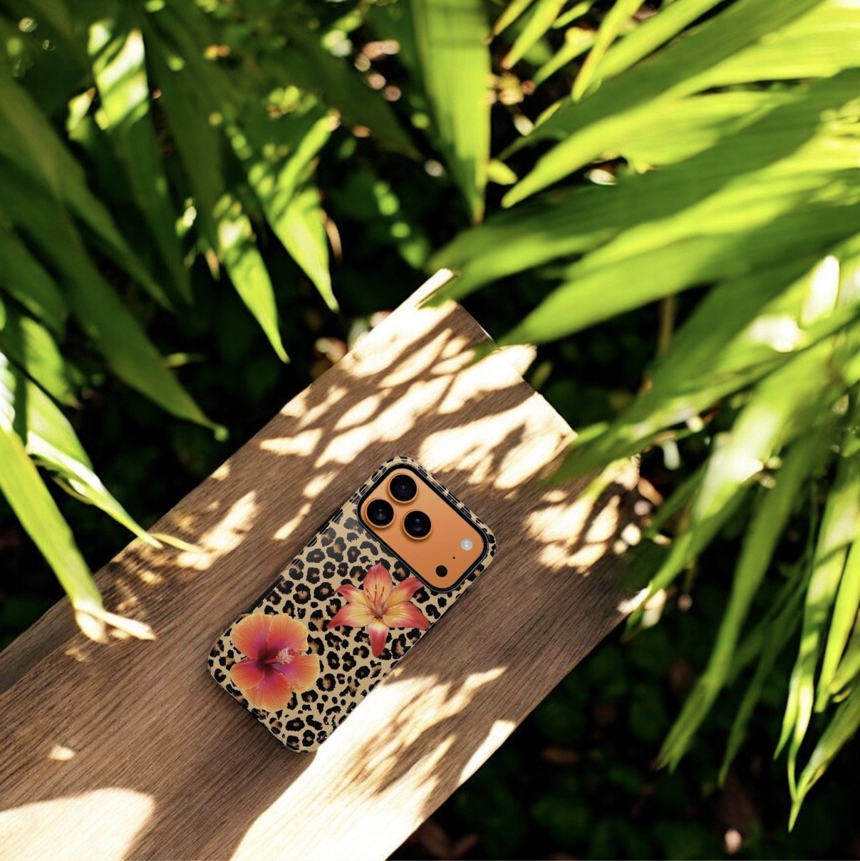 A smartphone with a leopard print case and floral design is placed on a wooden surface, surrounded by green plants and leaves, with sunlight casting shadows.