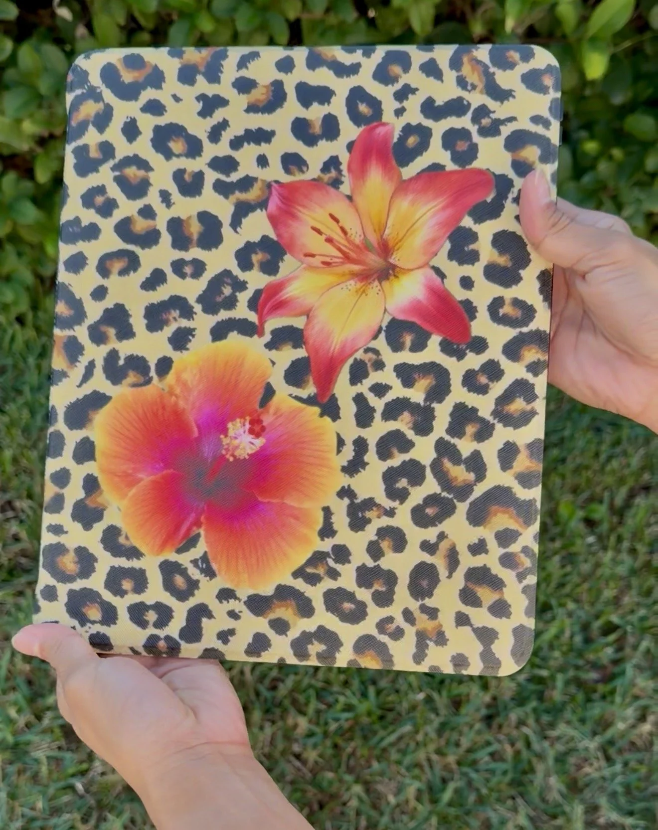 A square decorative item featuring a leopard print pattern with two large colorful flowers, one in yellow and pink, and the other in orange and pink, being held by two hands outdoors with greenery in the background.