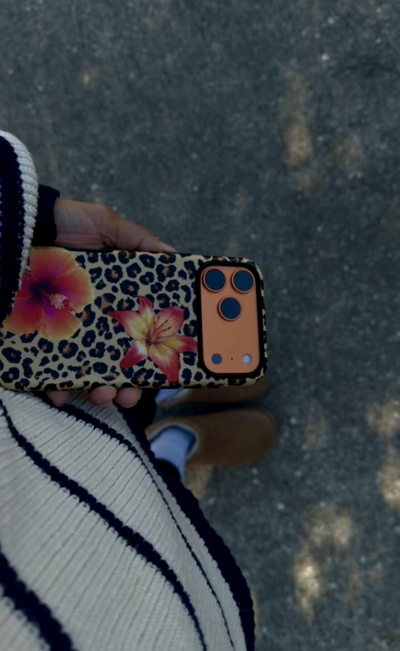 A person holding a smartphone with a floral, animal print phone case. The person is wearing a white sweater with navy stripes, and tan shoes. The ground is asphalt.