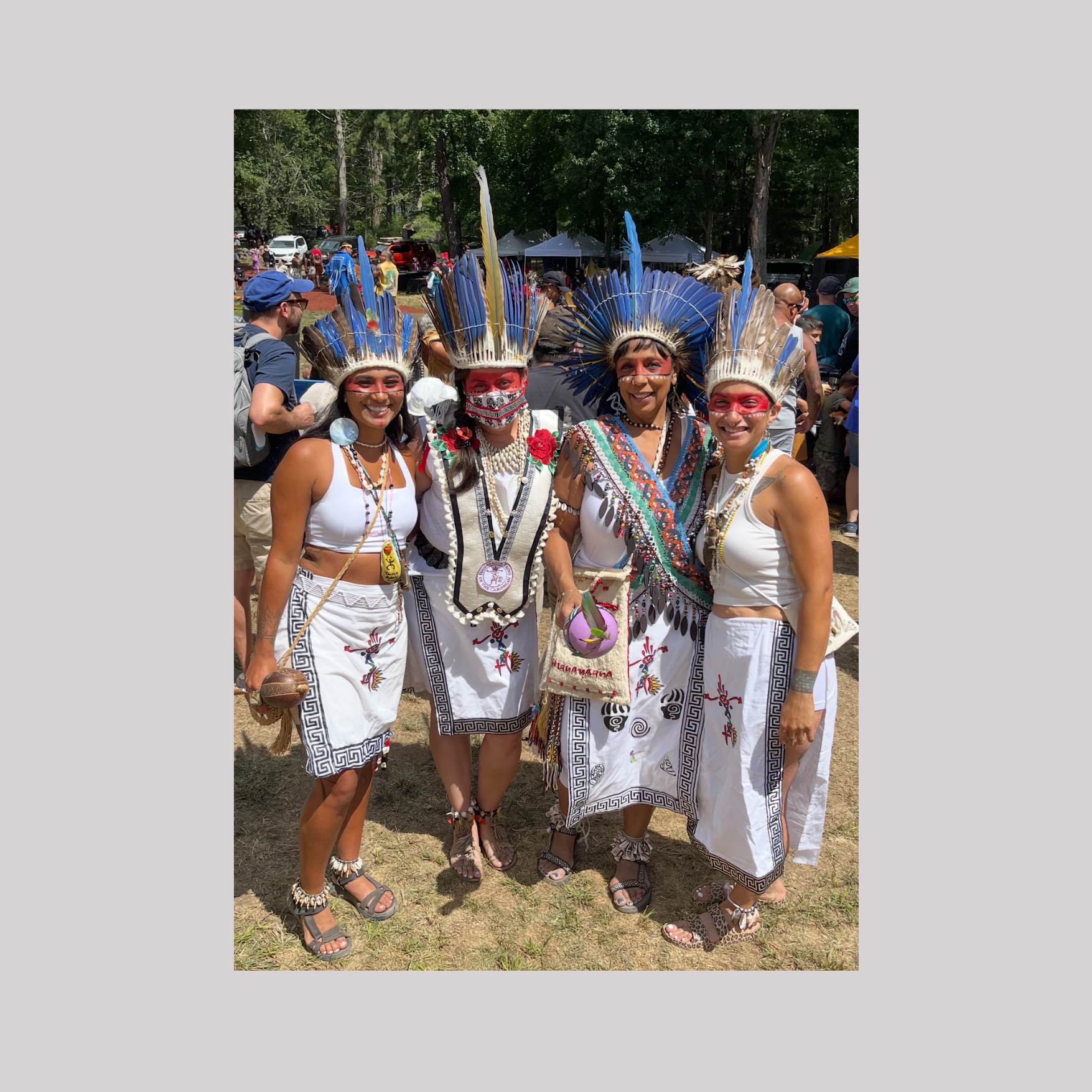Four women dressed in traditional Native American attire, wearing feathered headdresses, standing outdoors at a cultural festival.