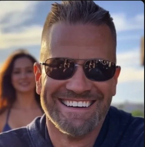 Close-up of a smiling man wearing sunglasses, with a woman in the background outdoors on a sunny day.