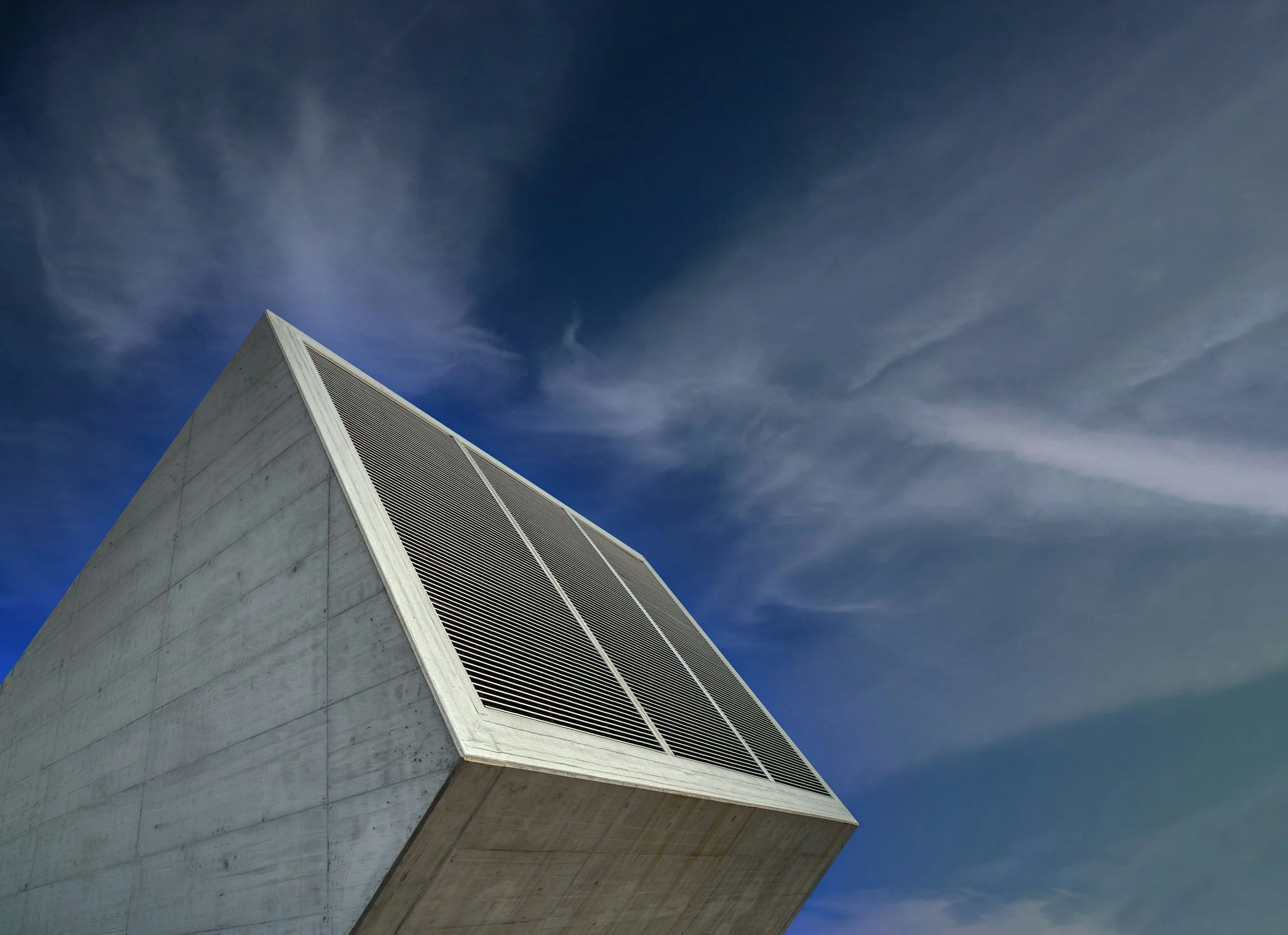 A modern concrete building with a tilted facade and large ventilation grid, set against a partly cloudy sky.