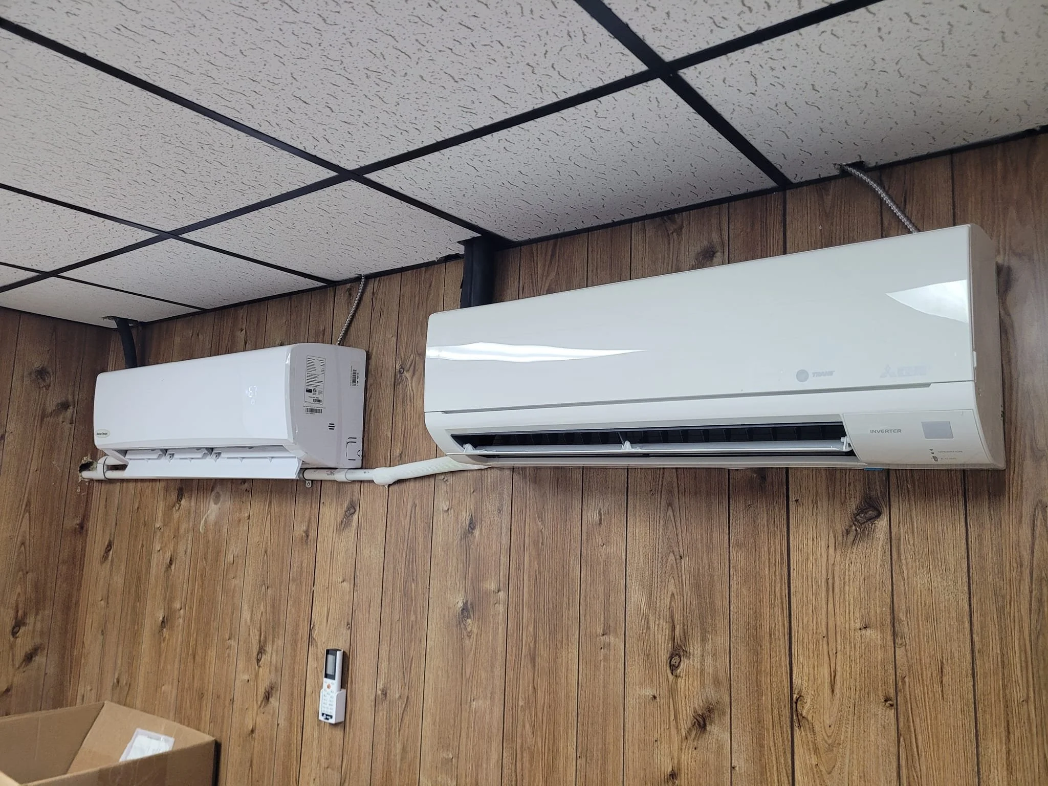 Two wall-mounted air conditioning units on a wooden wall, with a remote control below.