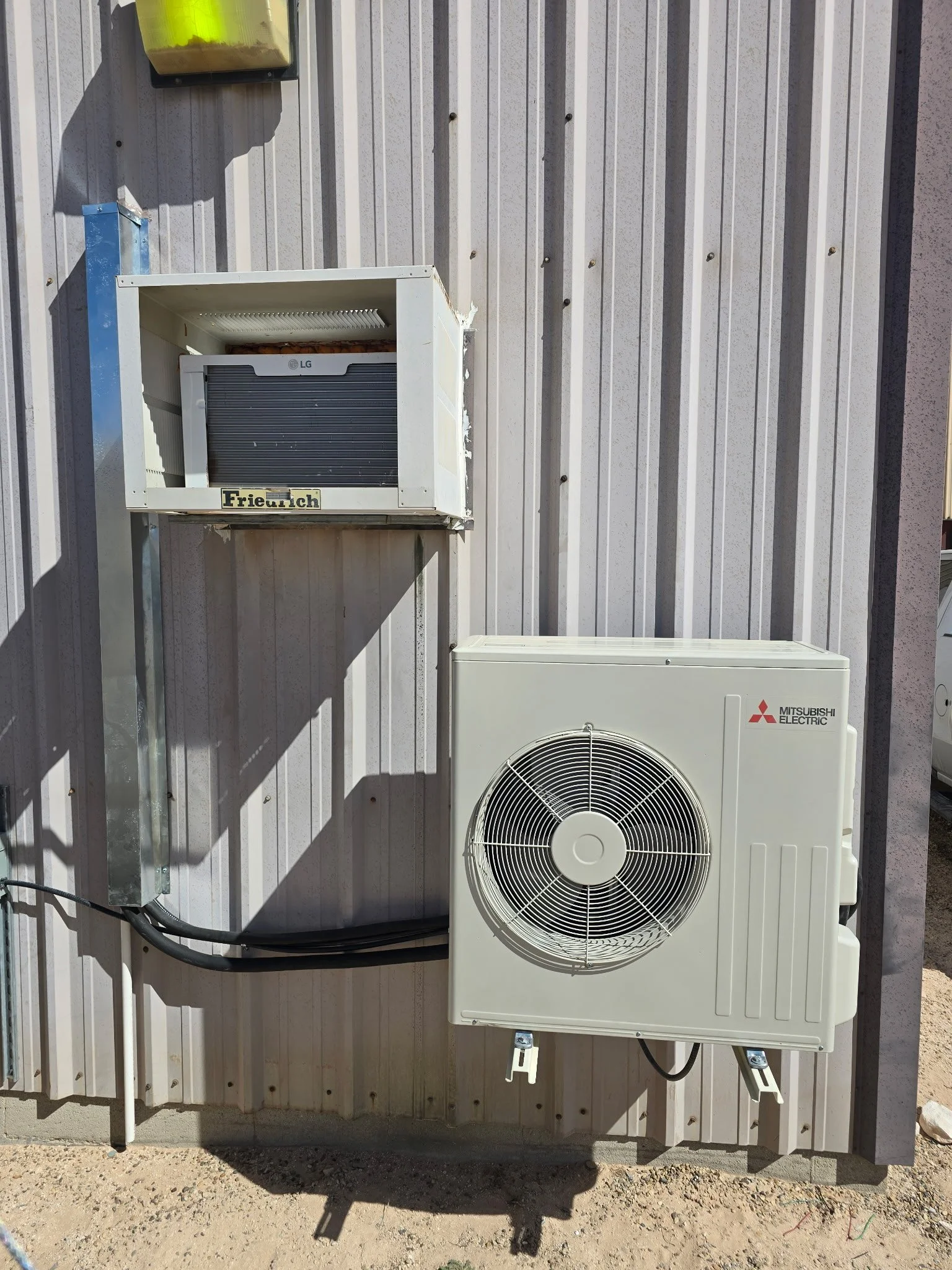 Outdoor view of an HVAC system with a Mitsubishi Electric unit and a smaller unit, mounted on a metal and siding wall.