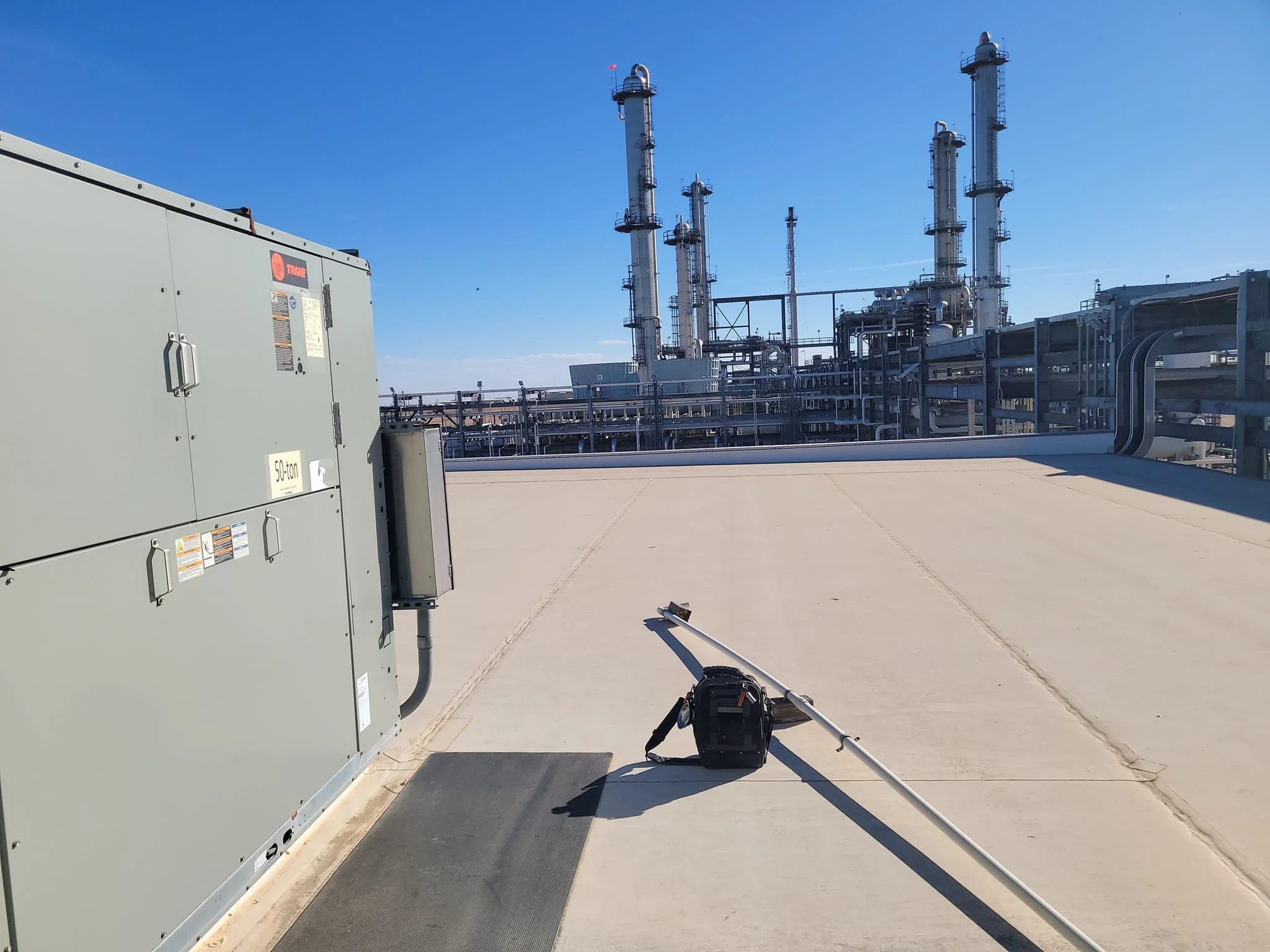 View of an industrial rooftop with large HVAC equipment, a black tool bag, and a long metal pole, with a refinery or chemical plant with multiple tall distillation columns and pipes under a clear blue sky in the background.