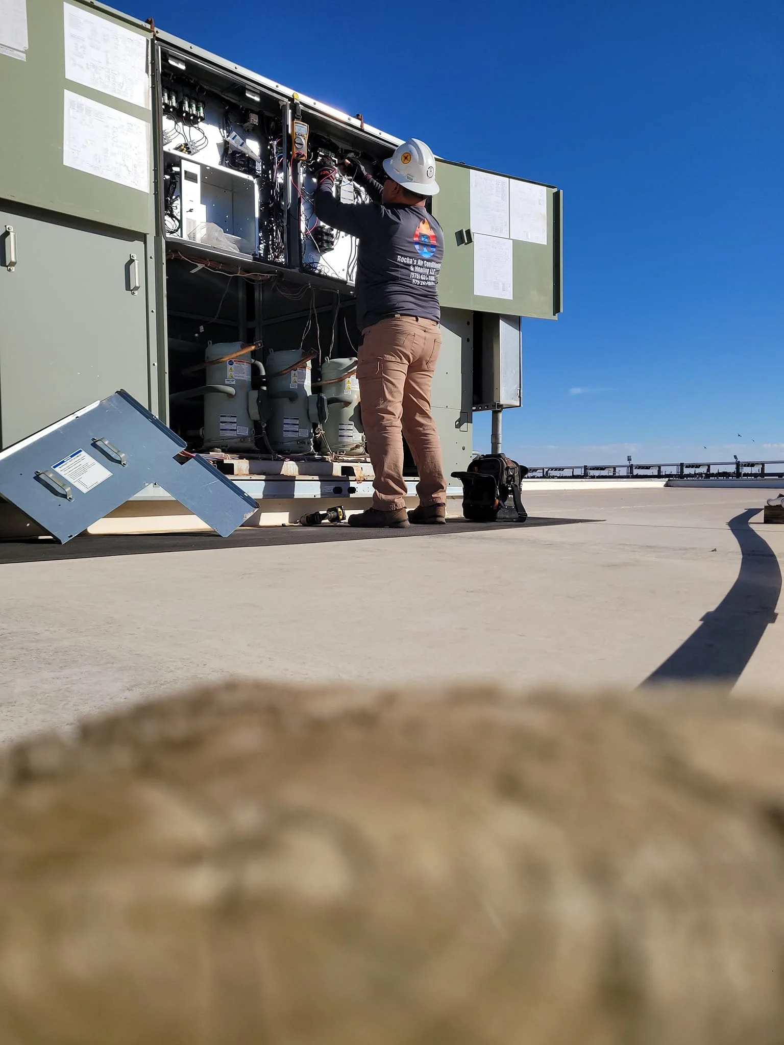 A technician wearing a hard hat and navy shirt working on electrical equipment outdoors on a cleared, flat rooftop under a clear blue sky.