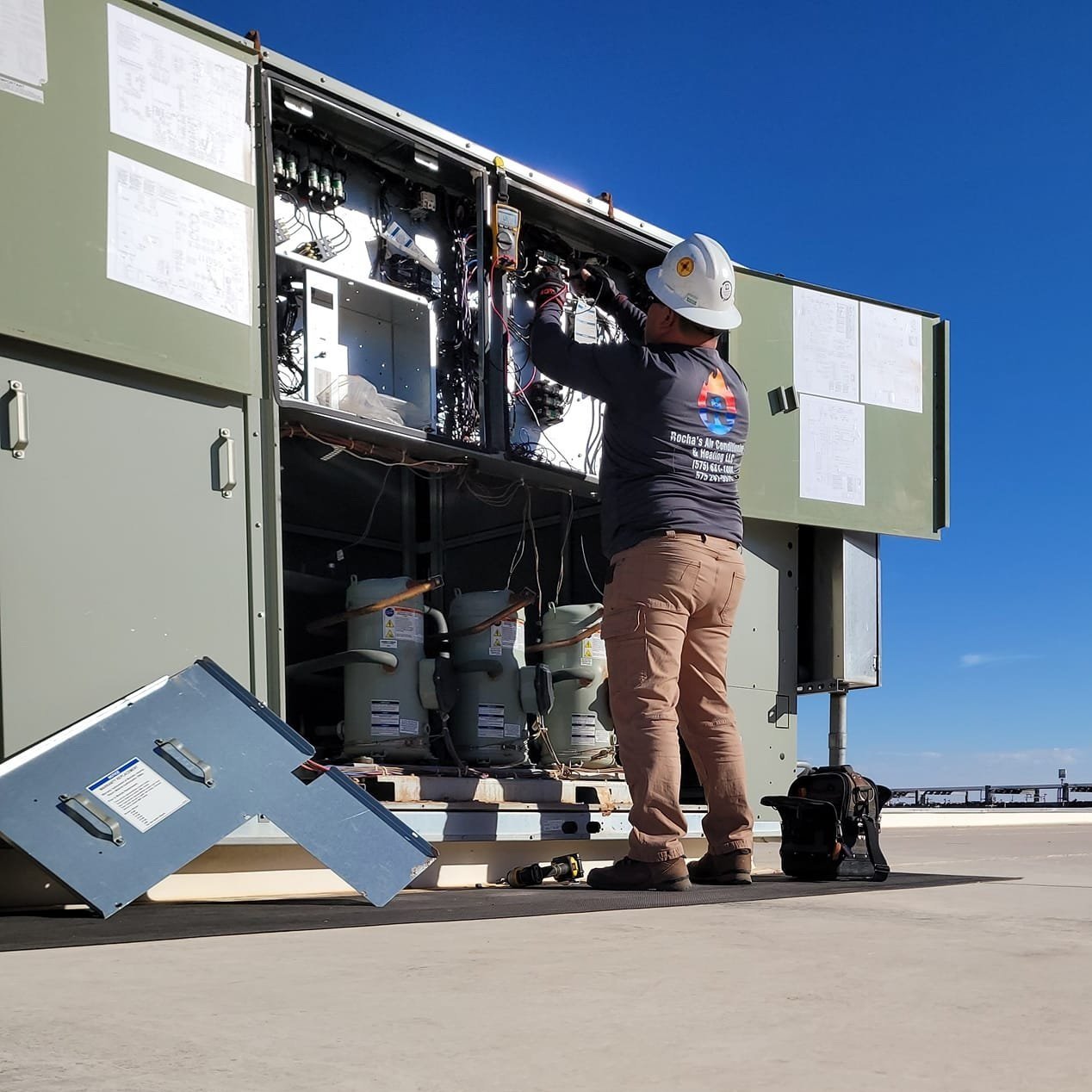 A worker in a hard hat and long sleeve shirt working inside an air conditioning unit on a rooftop solarpanel under a clear blue sky.
