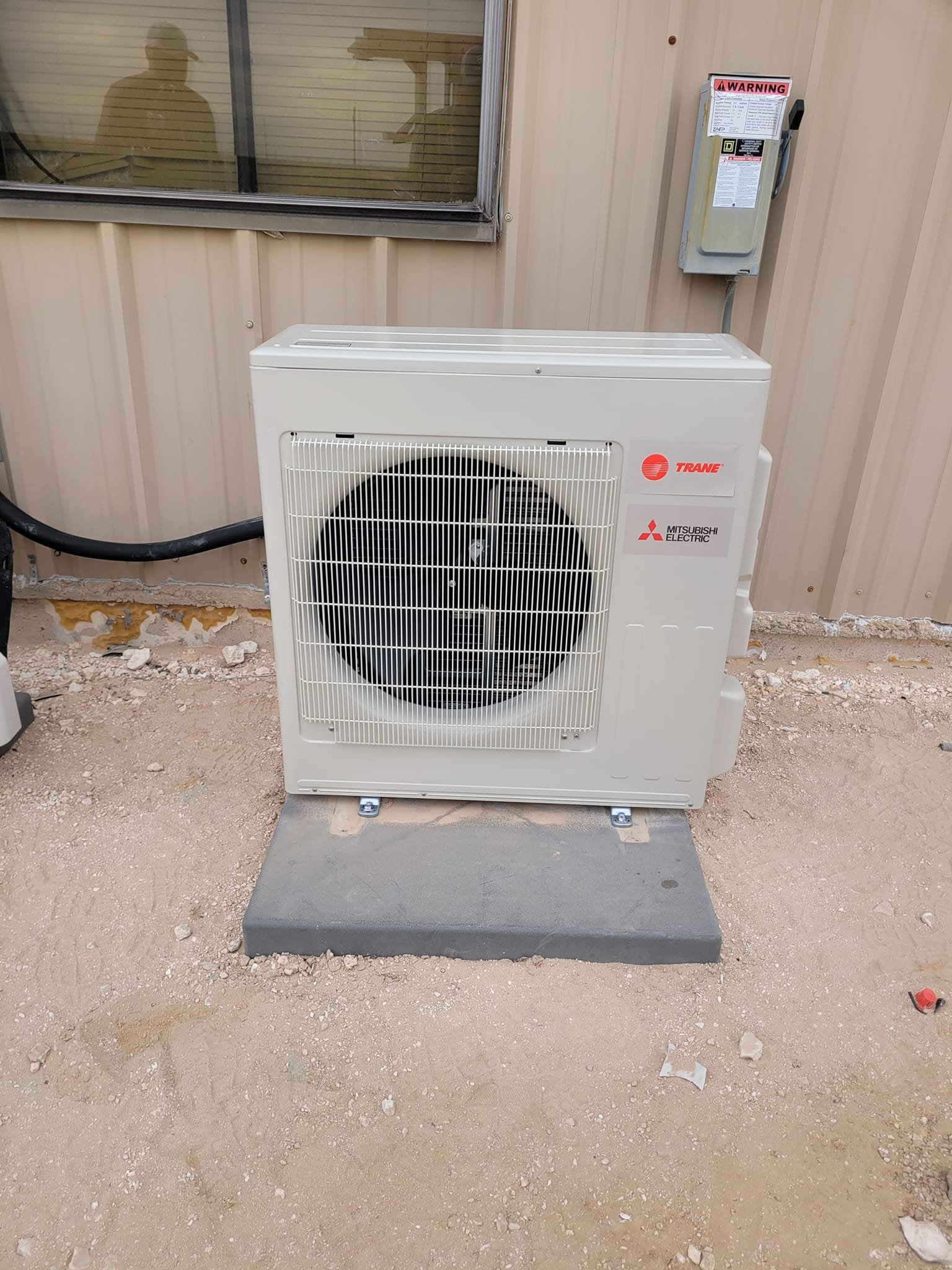 Outdoor air conditioning unit with Mitsubishi Electric and Trane logos, mounted on a concrete slab outside a building with siding, next to a window and an electrical box.